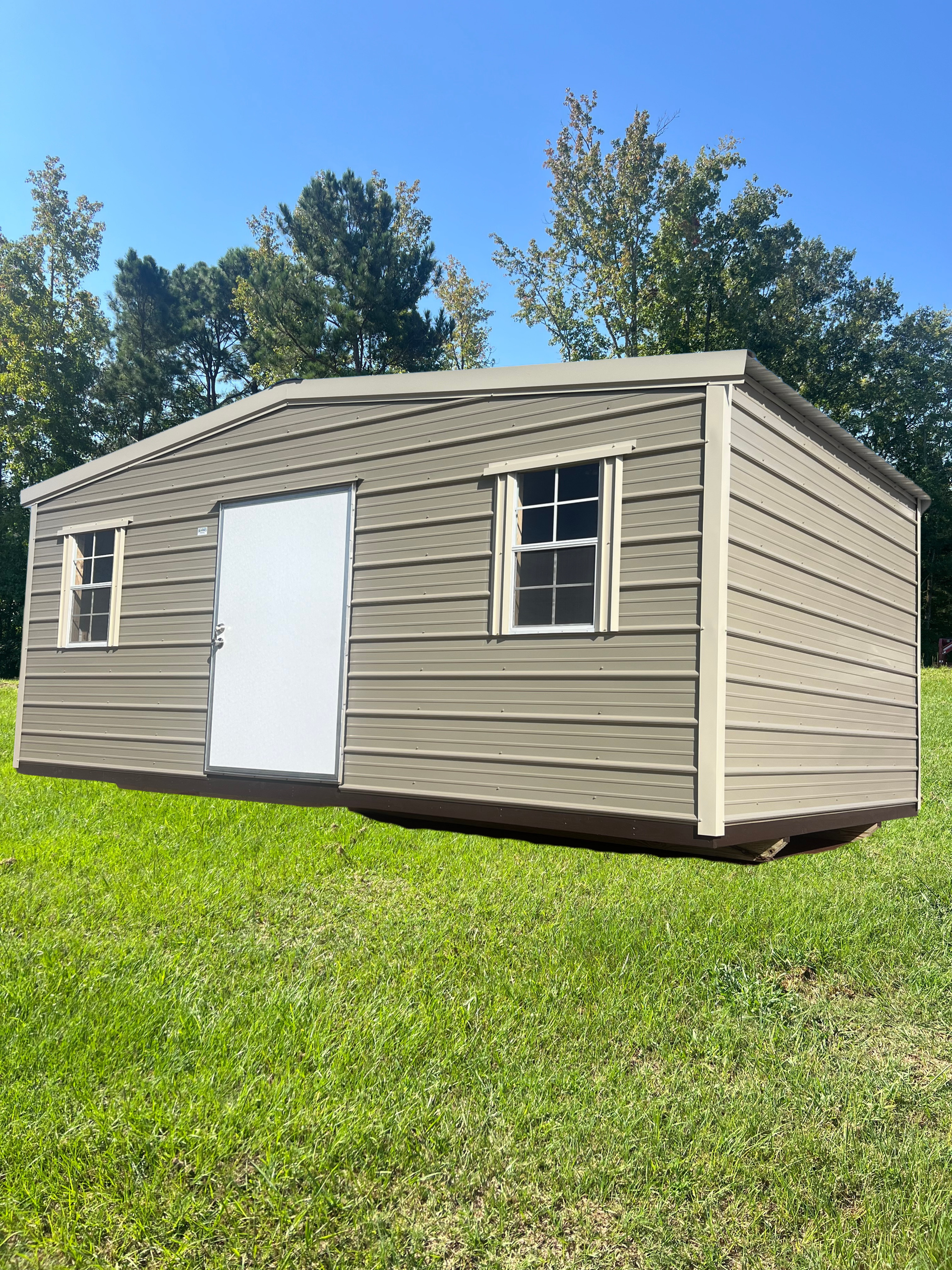 Tan shed with white door and windows on green grass under a blue sky.