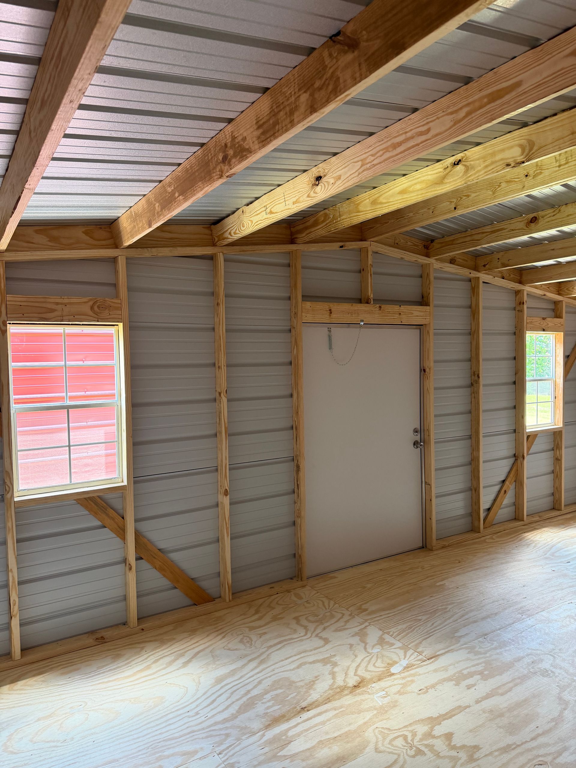 Interior of unfinished wooden shed with exposed beams, gray walls, and a white door centered between two windows.