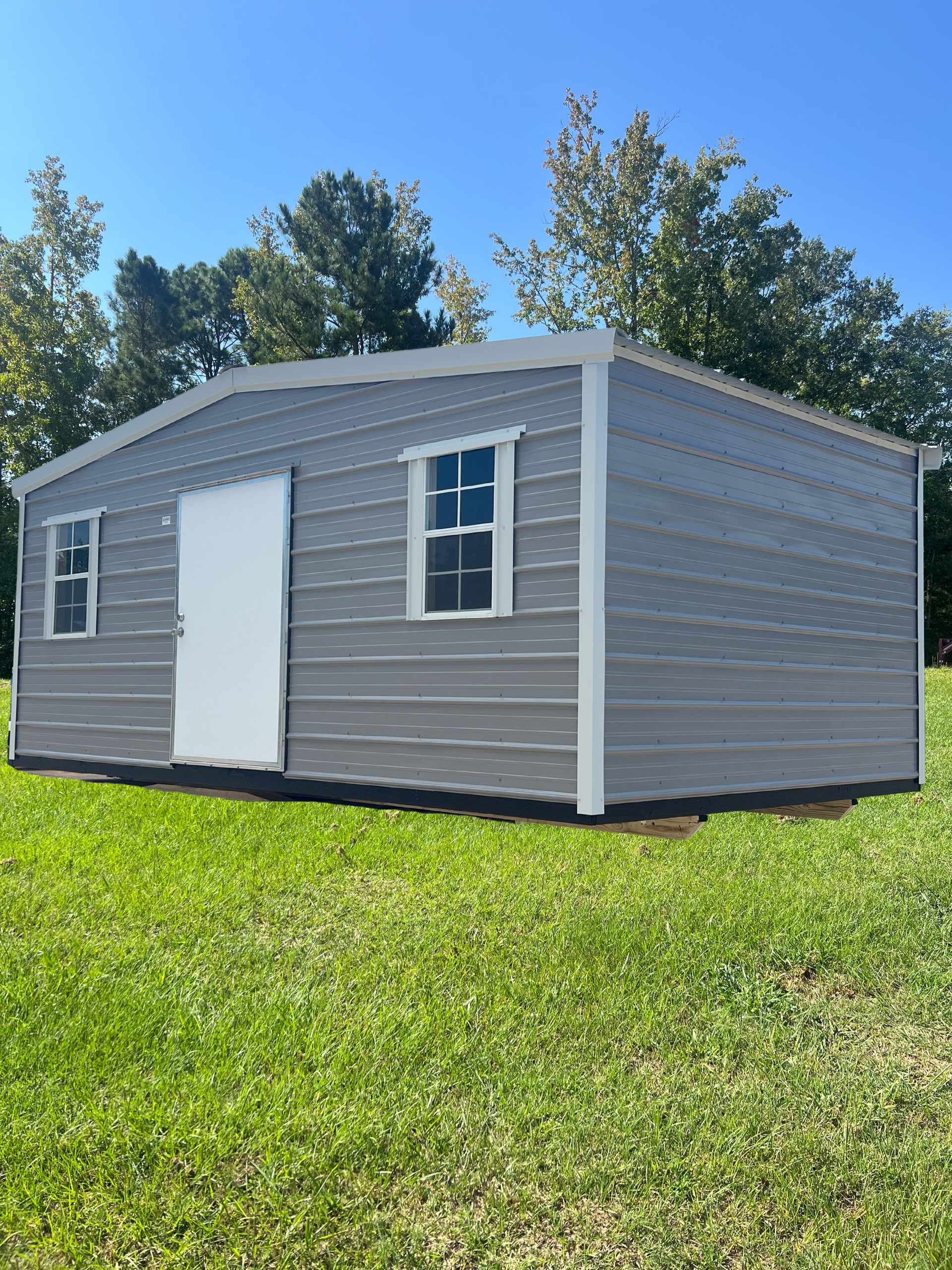 Small gray shed with white door and window sitting on a grassy lawn under a blue sky.