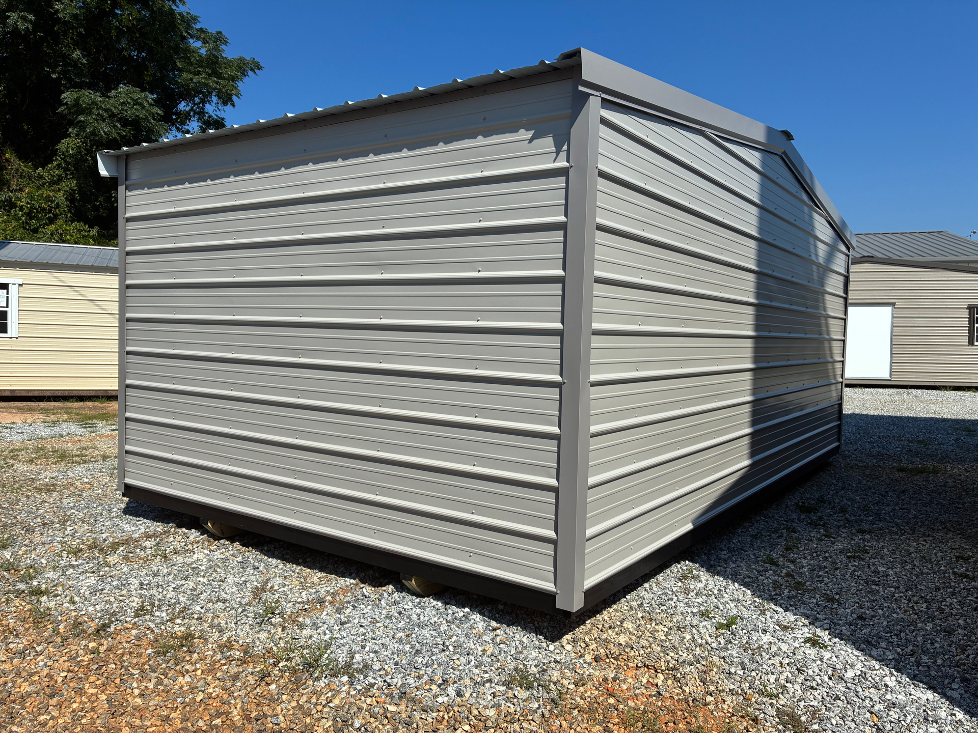 Gray metal storage shed with horizontal siding on a gravel lot under a clear blue sky