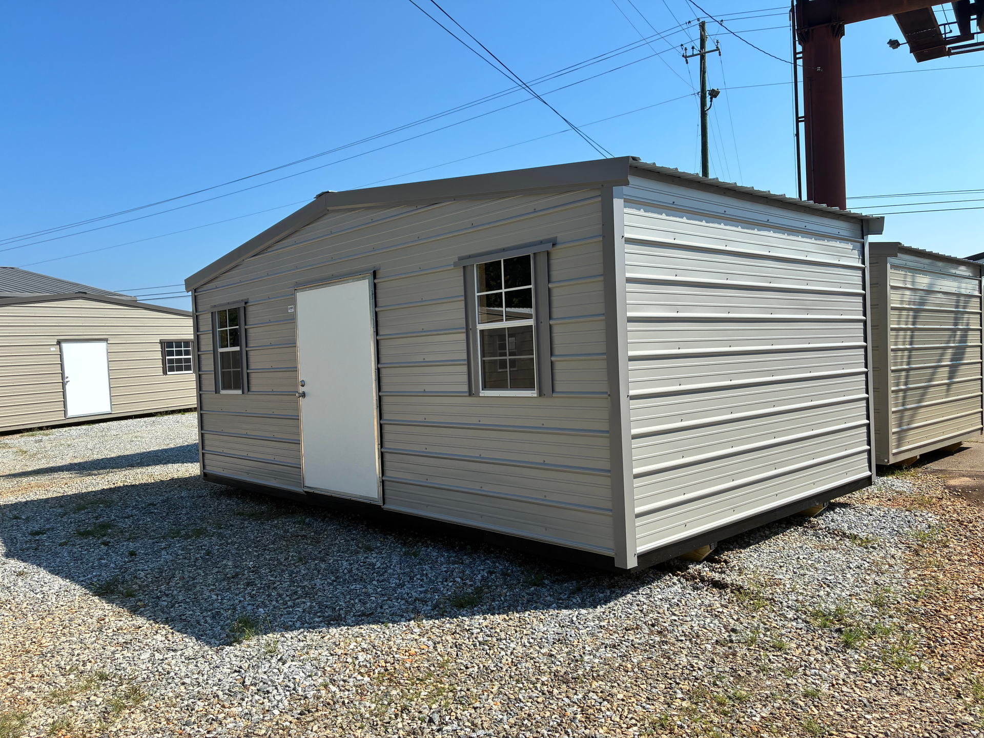 Small beige storage shed with white door and windows on a gravel lot under a blue sky