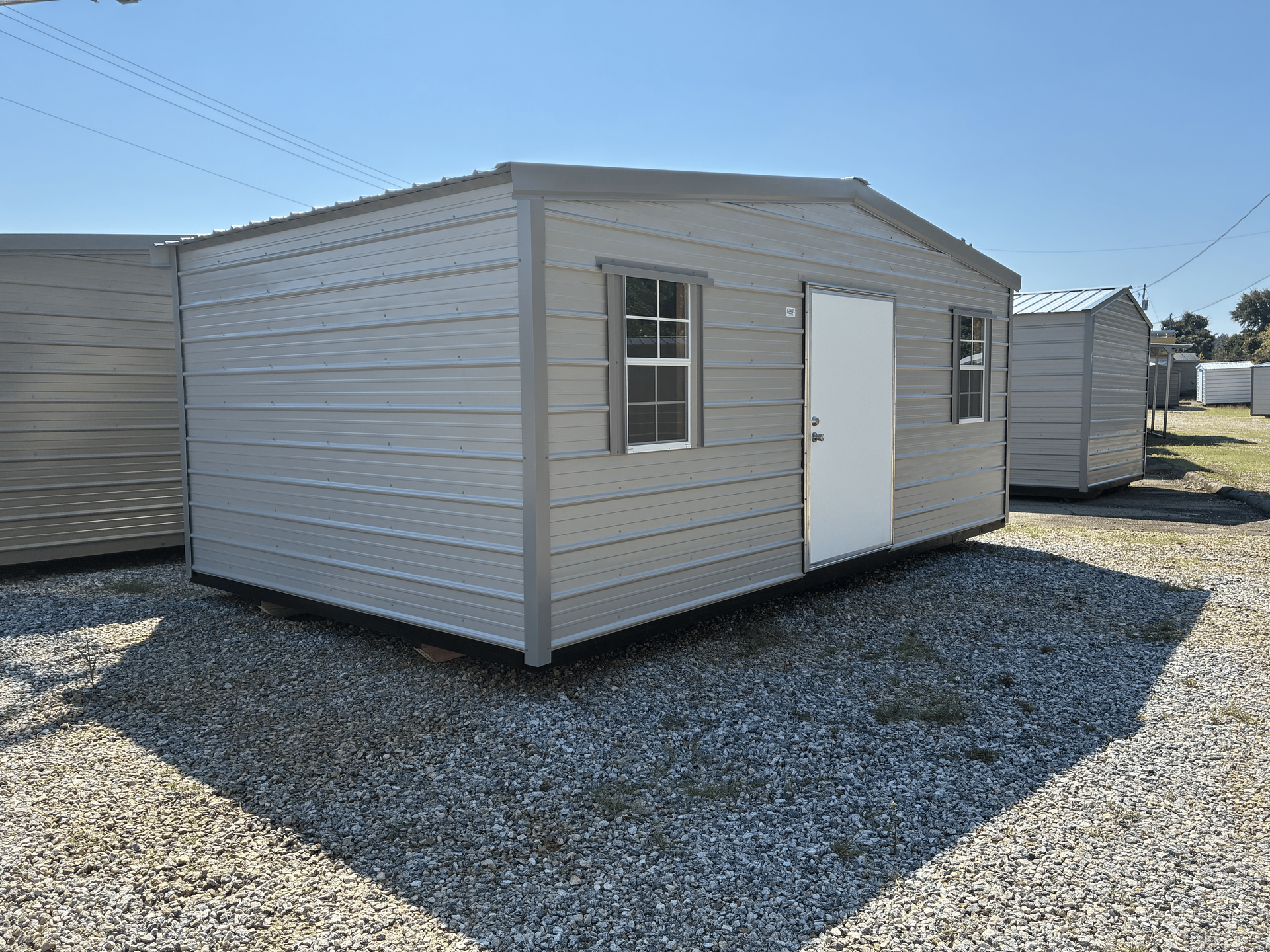 Small beige metal shed with white door and window on a gravel lot under a clear blue sky