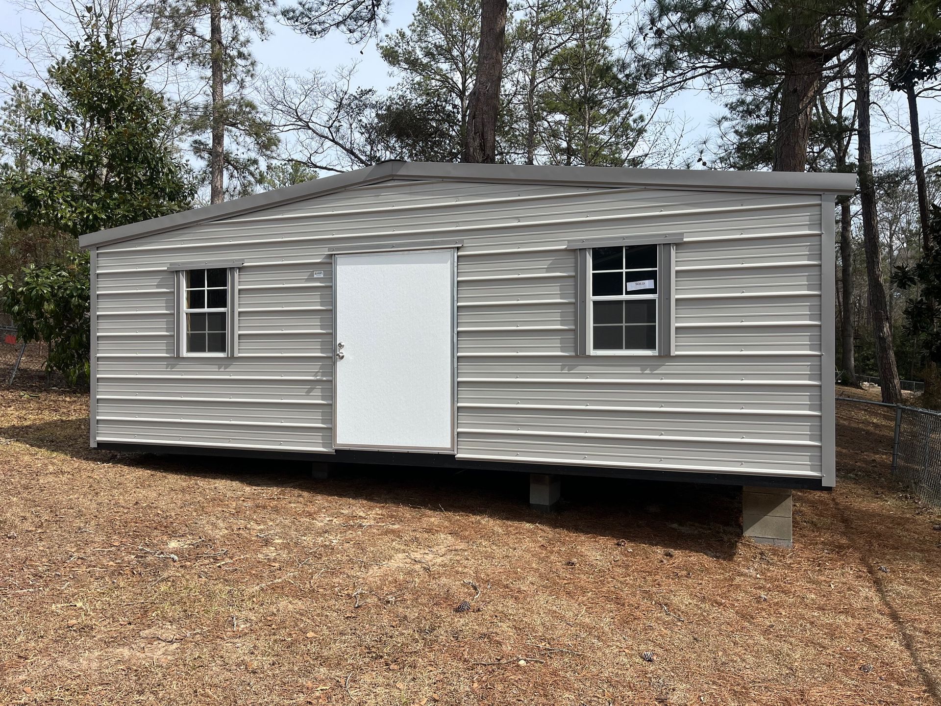 Gray metal storage shed with a white door and two windows, set on concrete blocks.