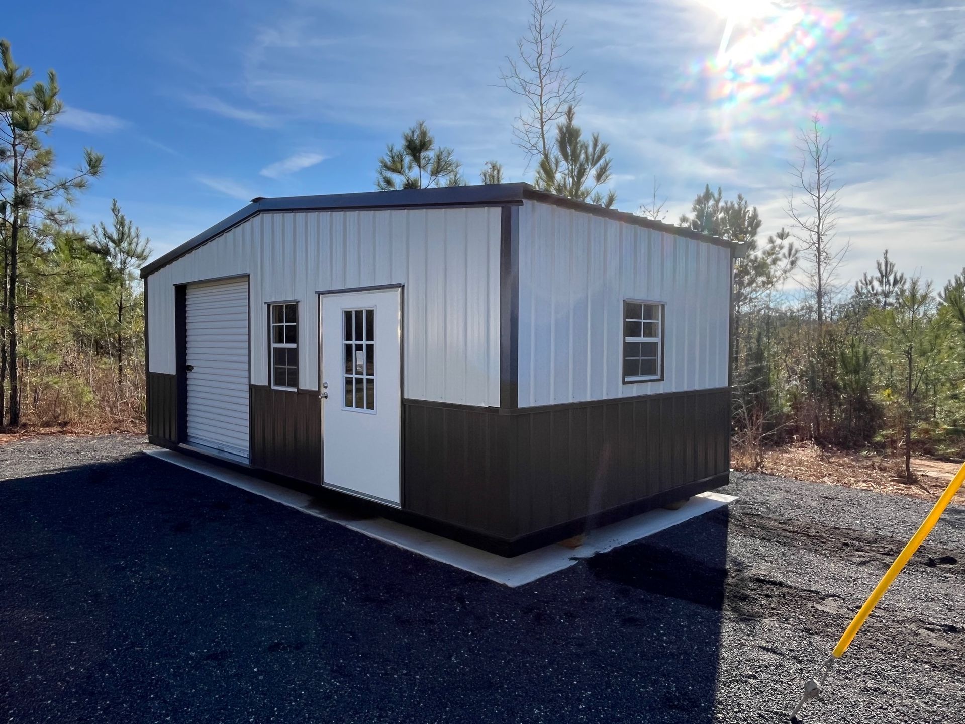 Metal shed with white and brown walls, garage door, entry door, and small windows, outdoors on gravel.
