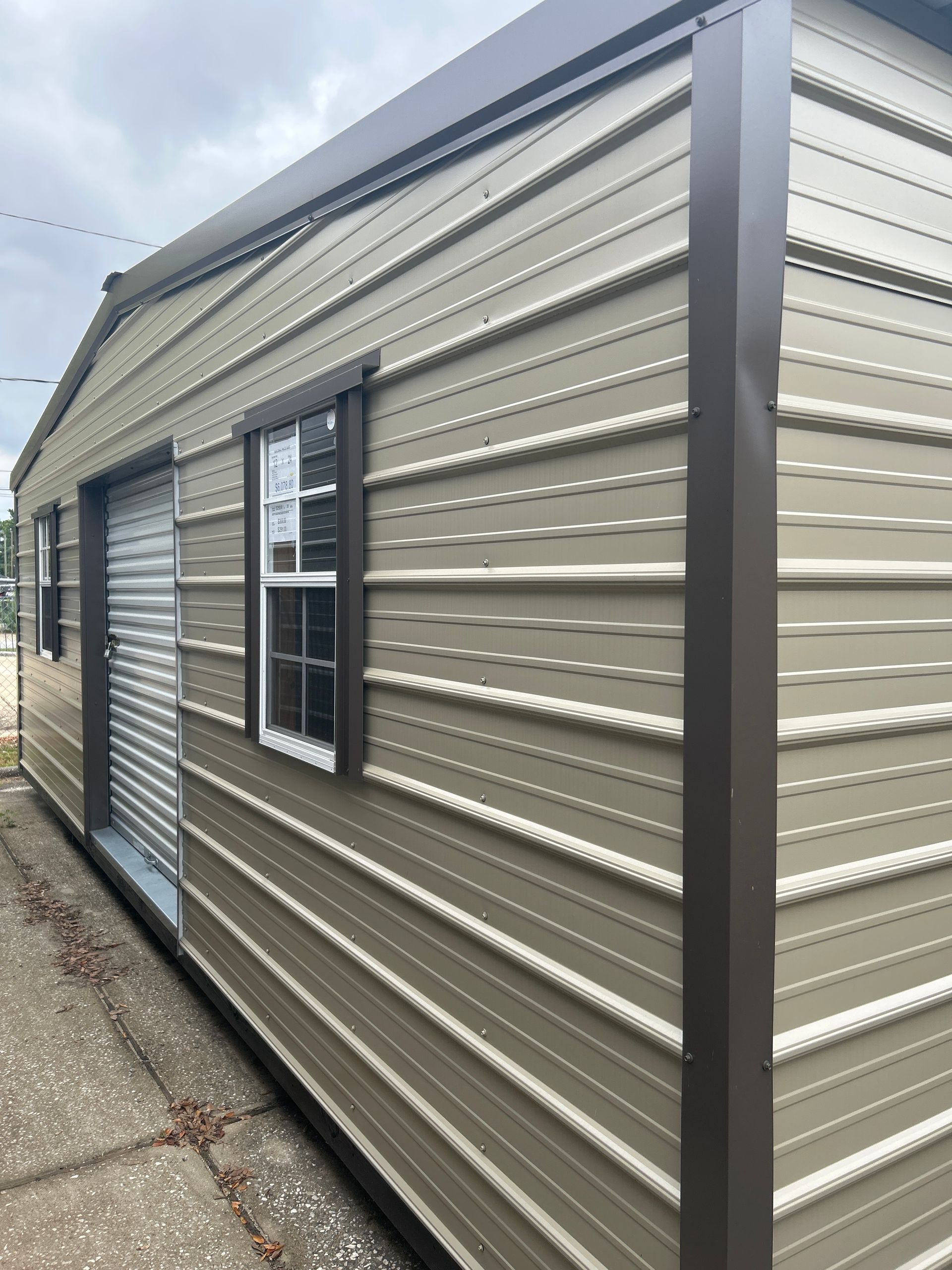 Tan metal shed with brown trim, a roll-up door, and a window, outside on a gray sidewalk.
