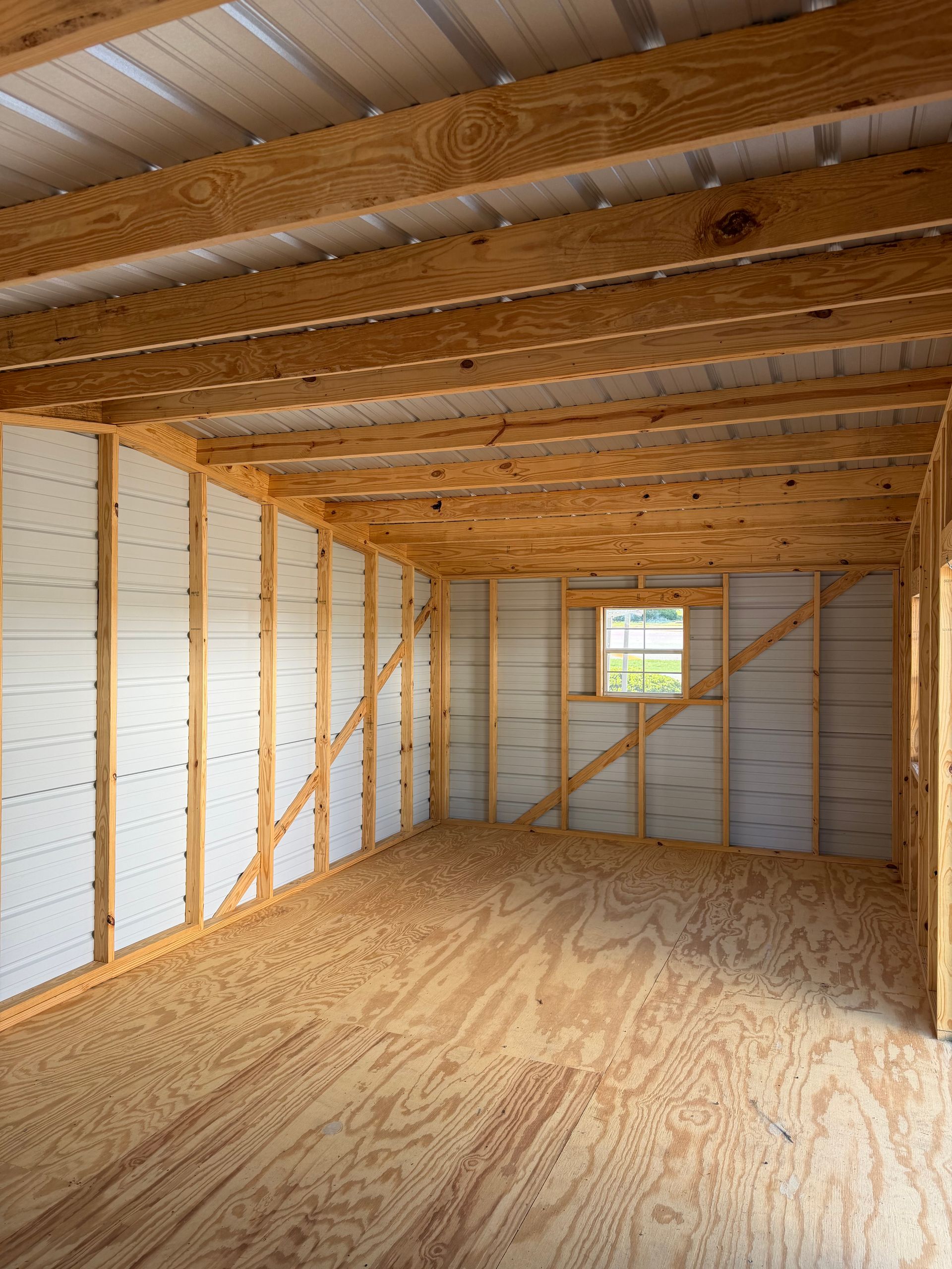 Empty unfinished room with exposed wooden beams, plywood floor, insulated walls, and a small window.