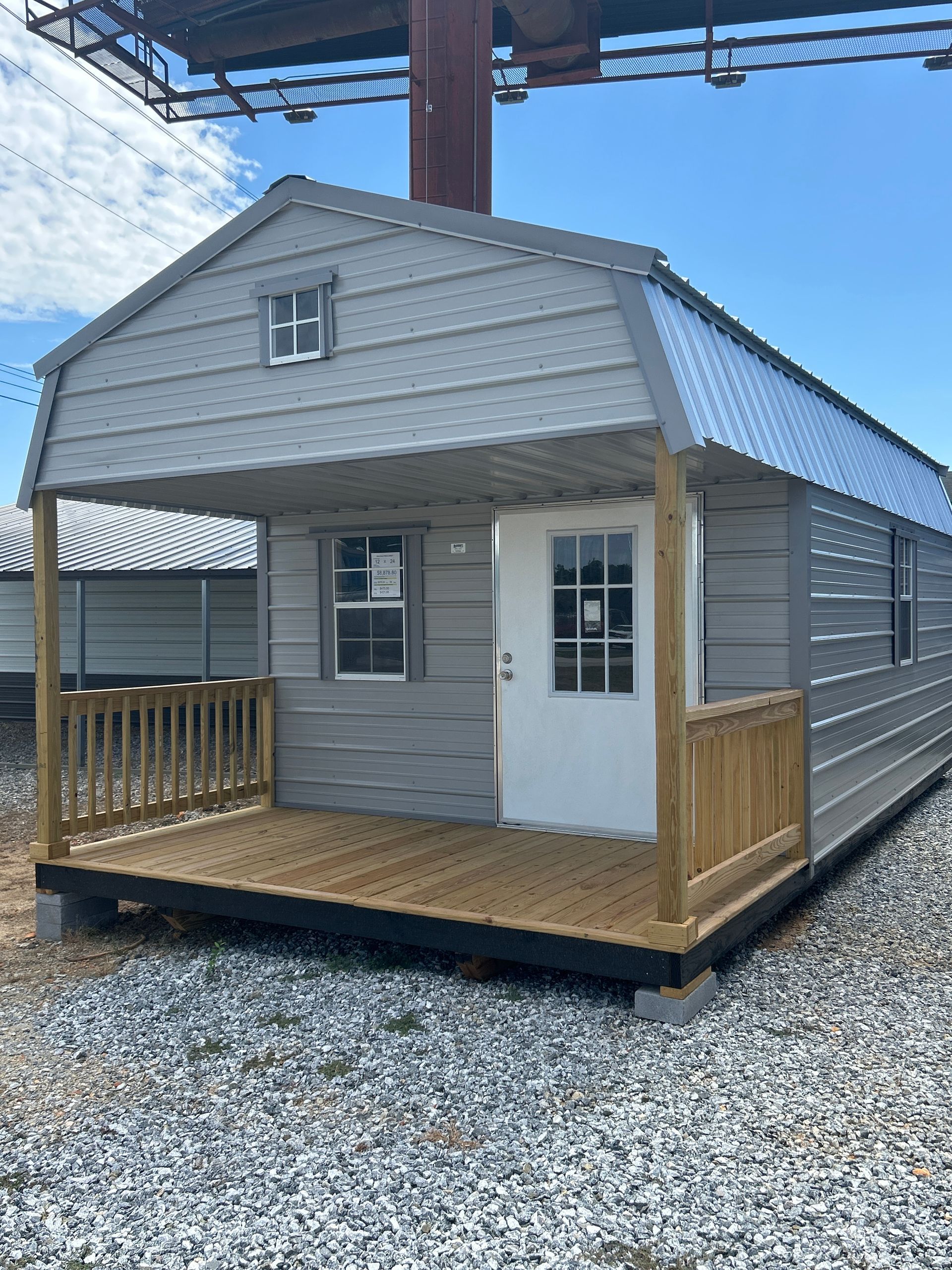 Gray cabin with porch, gray siding, white door, blue roof, set on gravel.