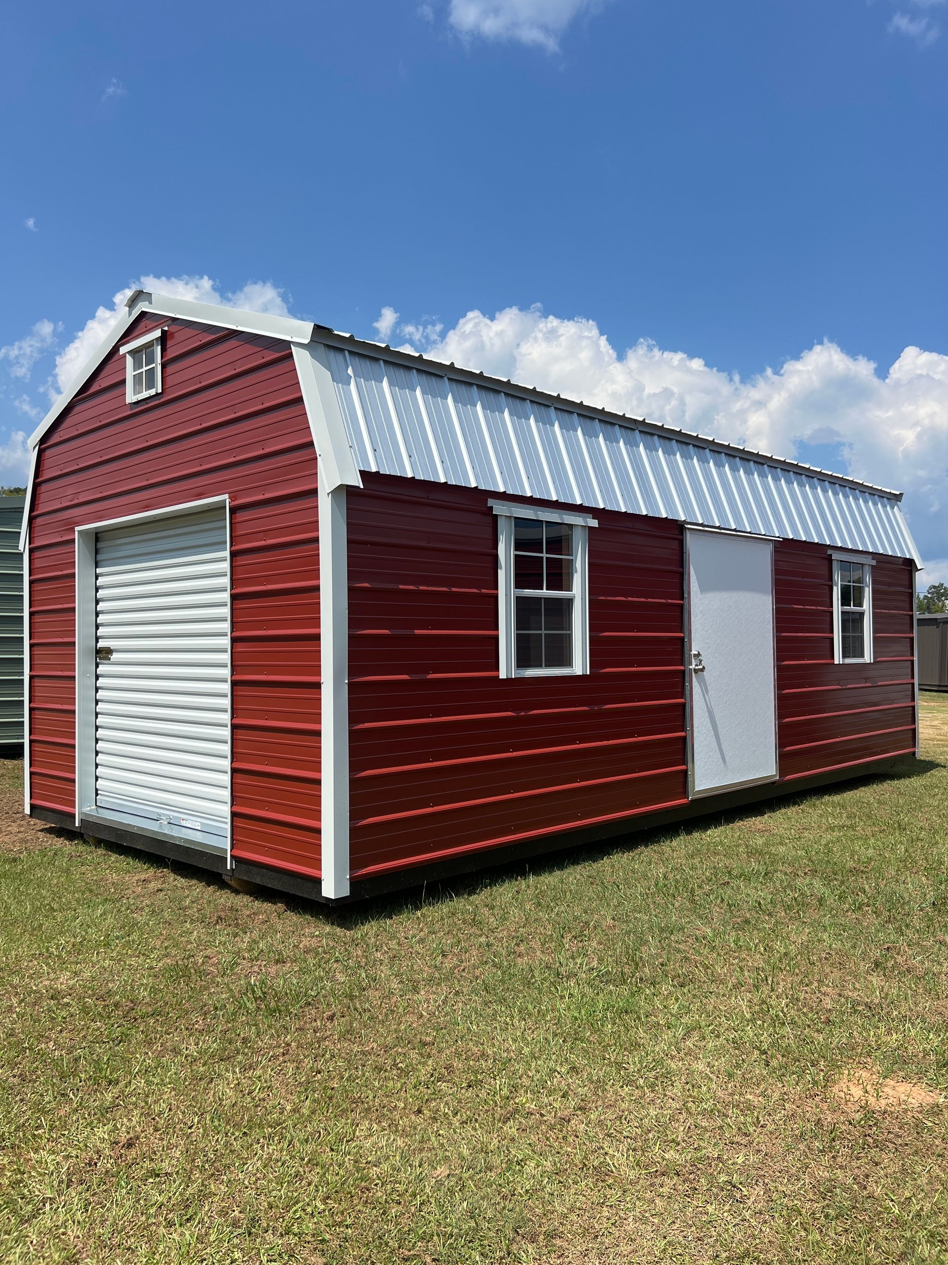 Red barn-style shed with white garage door, white door, and windows, set on a green lawn under a blue sky.