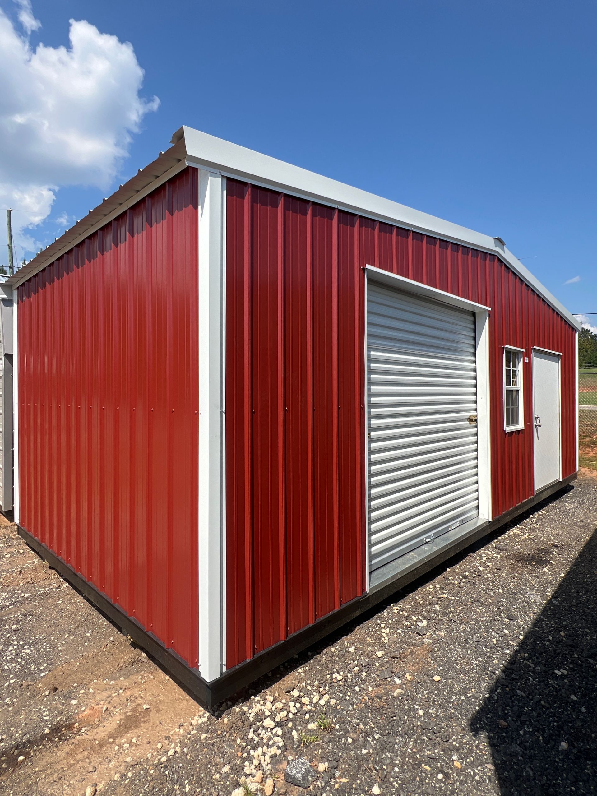 Red metal storage building with white trim and roll-up door, on gravel under a blue sky.
