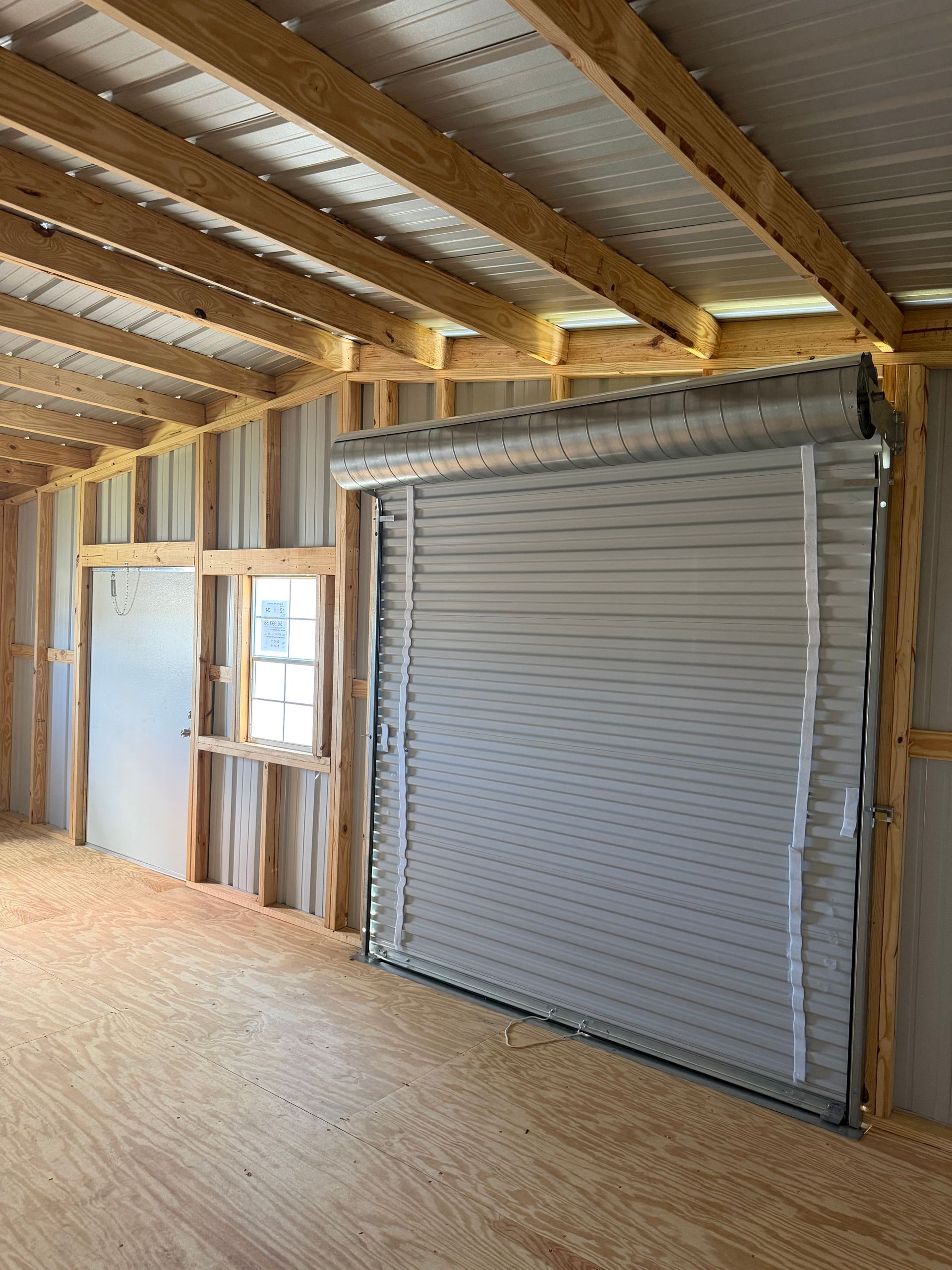 Interior of unfinished building with exposed beams, plywood walls, and a large gray roll-up door