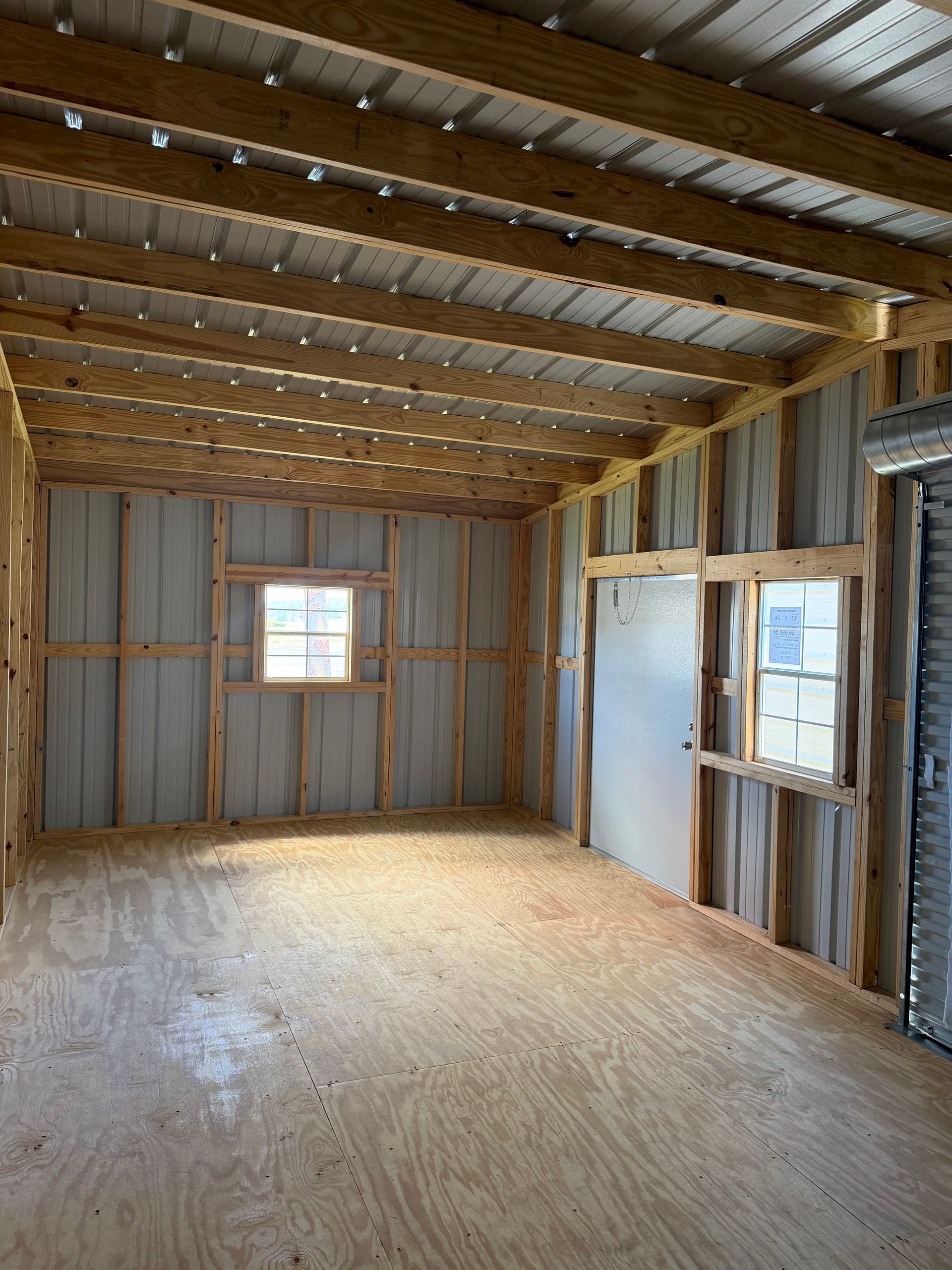 Empty unfinished room with exposed wood framing, plywood floor, and two small windows.