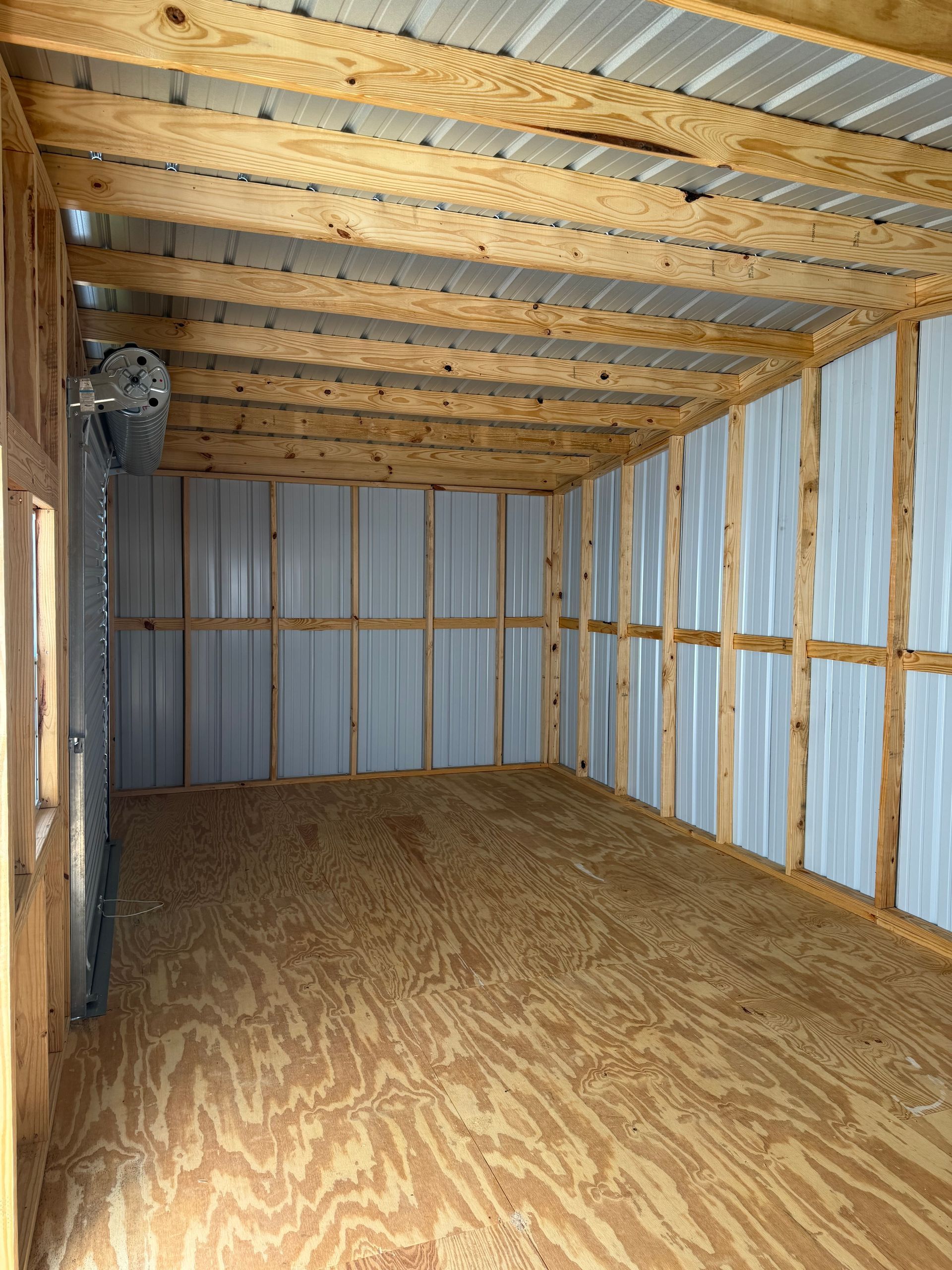 Empty unfinished shed interior with plywood floor, exposed wood framing, and corrugated metal walls