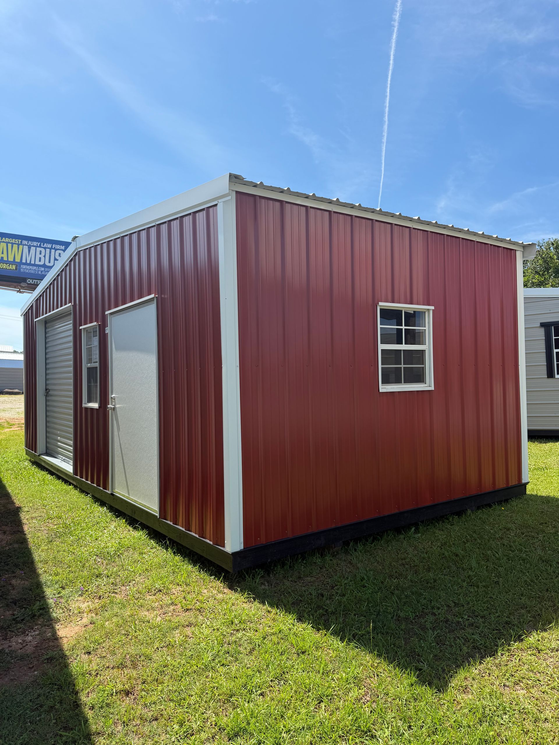 Red portable storage building with white trim and small windows sitting on grass under a blue sky