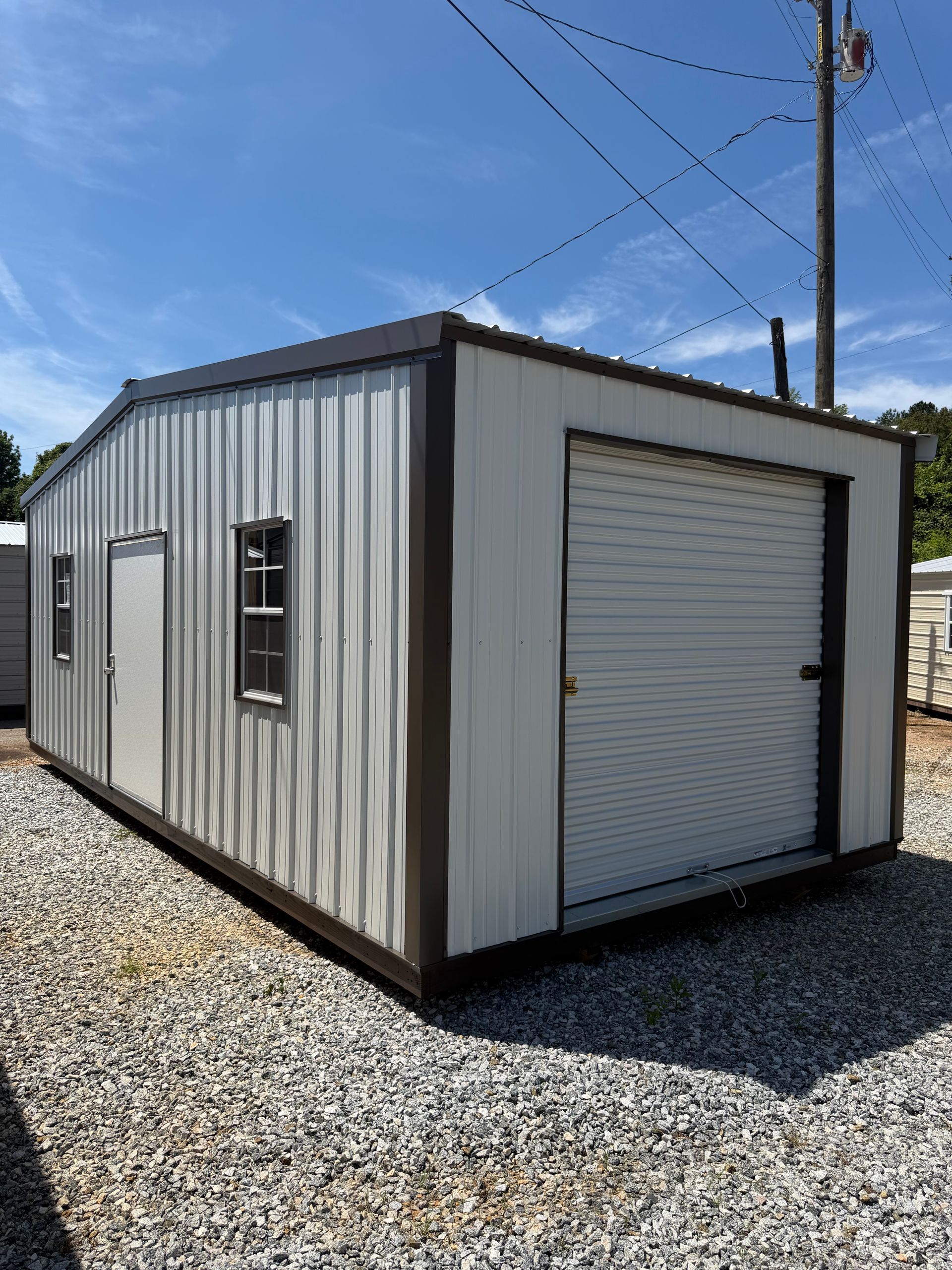 Small gray shed with a white door and windows sitting on a grassy lawn under a blue sky.