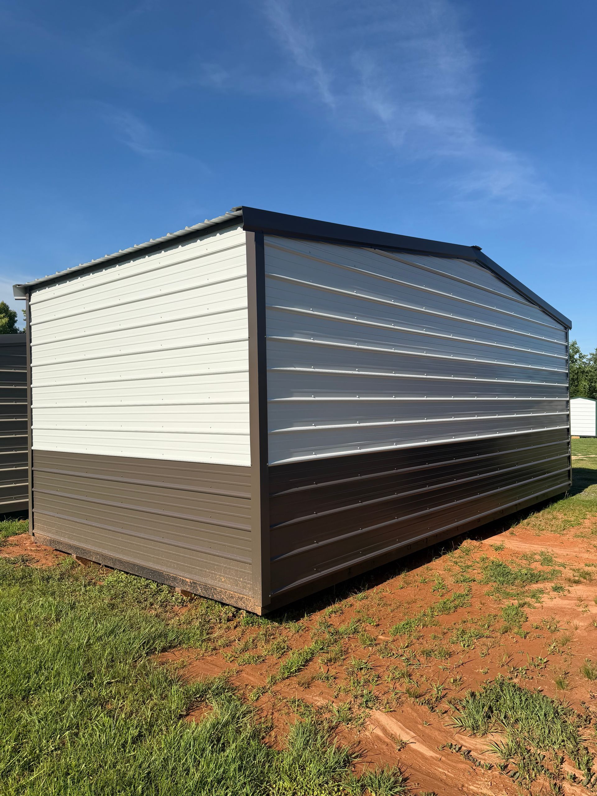 Small white-and-brown metal shed with a dark roof on a grassy dirt lot under a blue sky.
