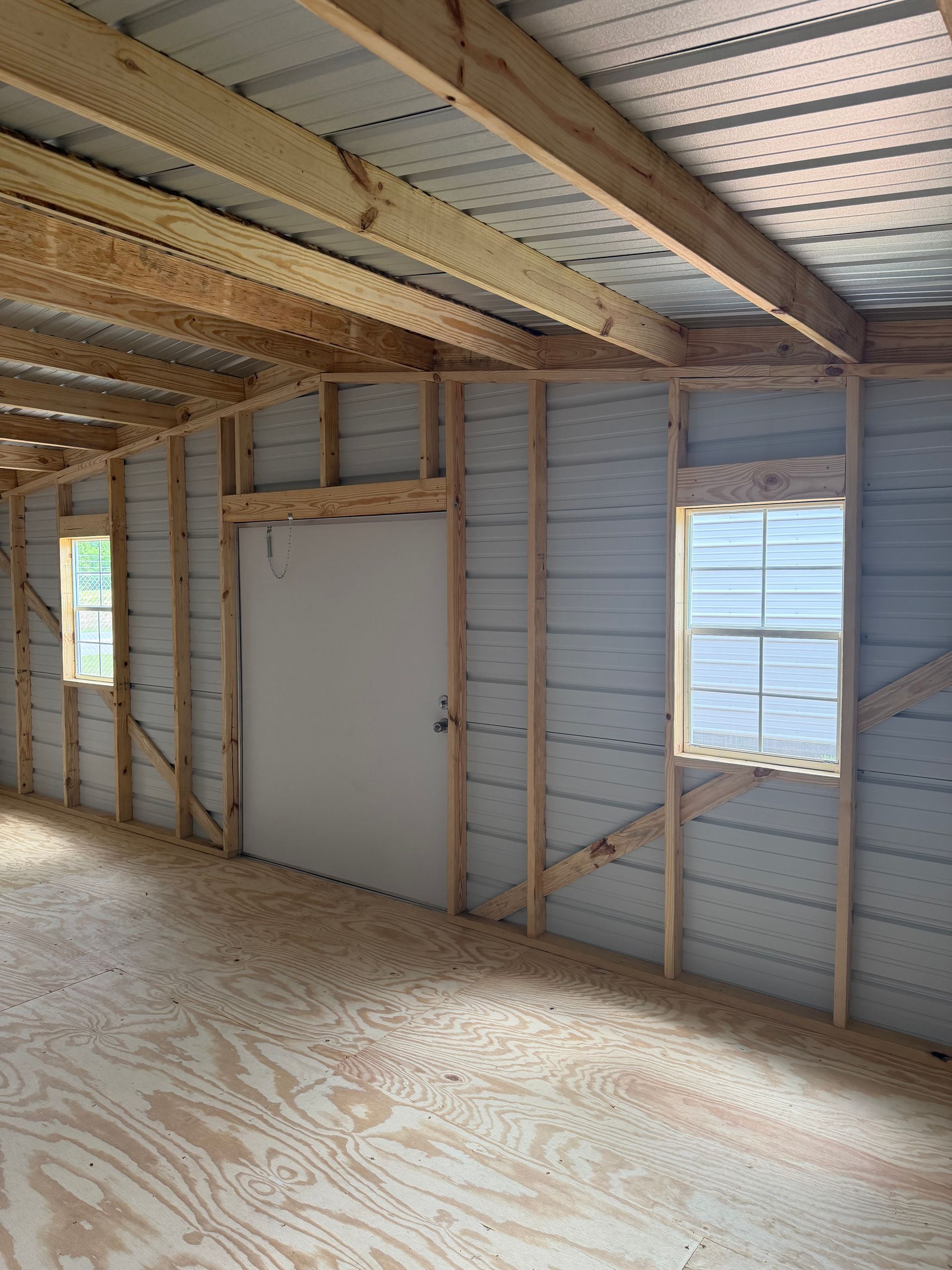 Interior of an unfinished room with exposed studs, plywood floor, white door, and small window.