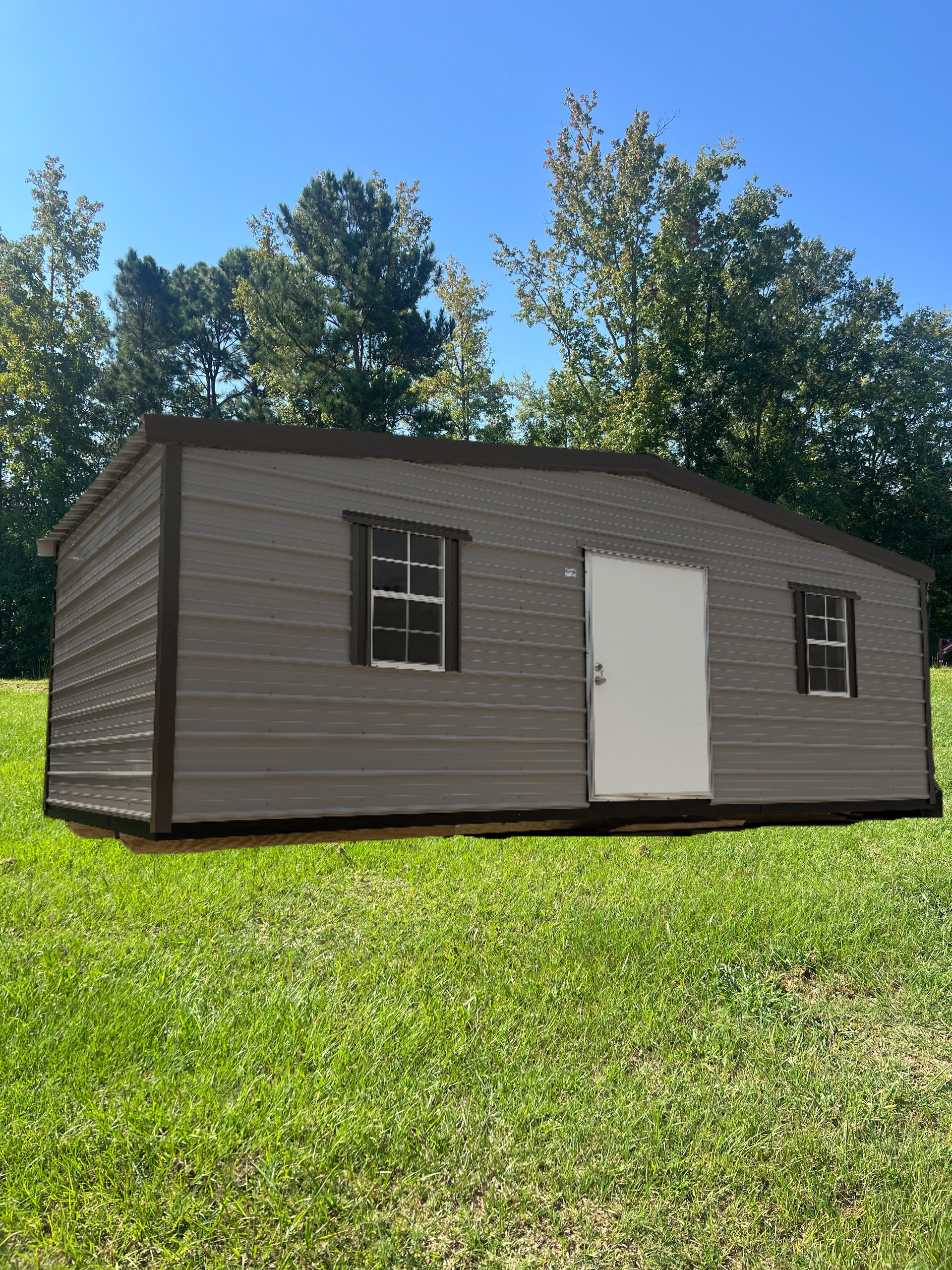 Small brown shed with white door and windows on a grassy lawn under a blue sky.