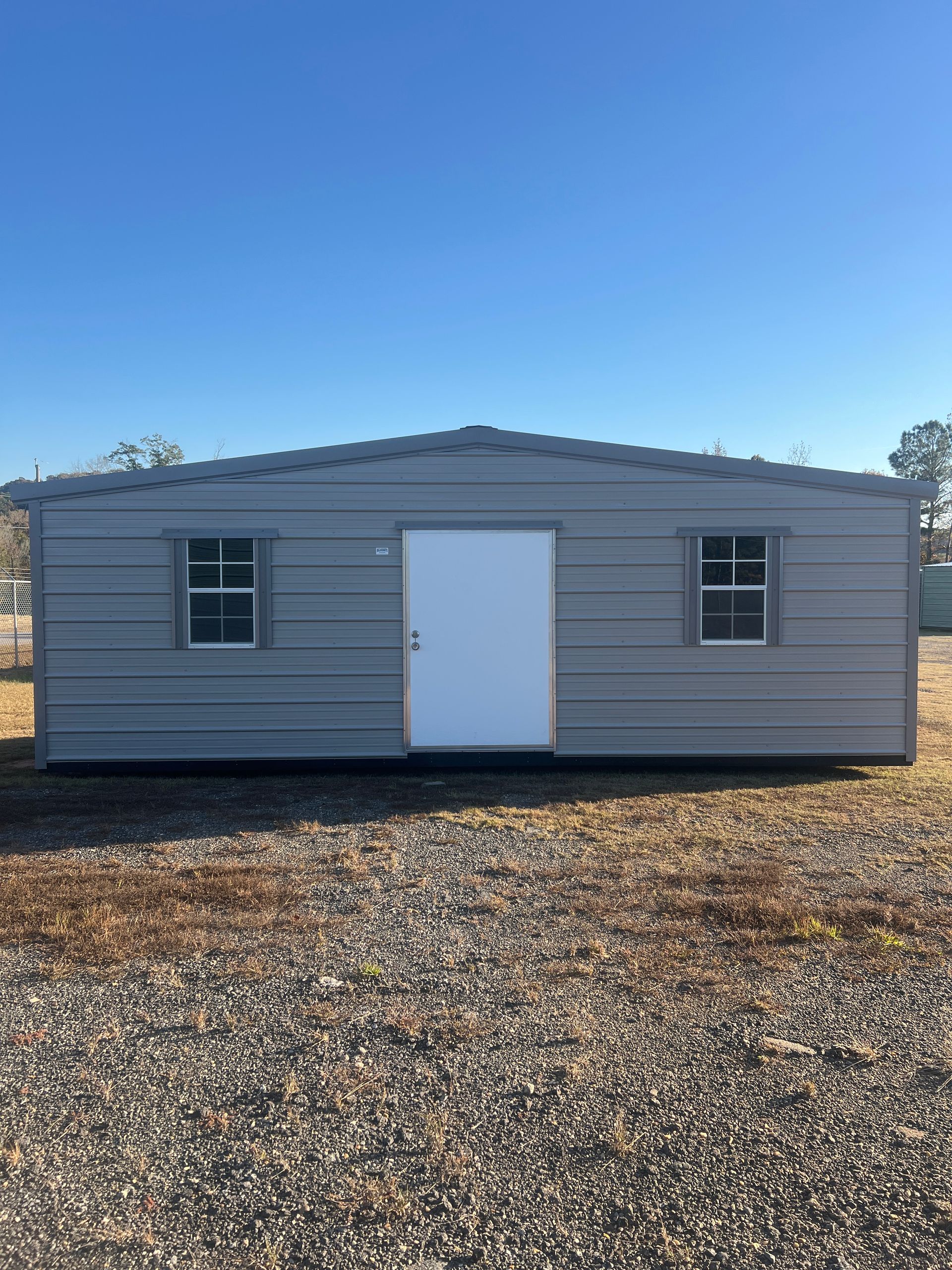 Tan metal building with white door, windows, and brown trim, set on pavement, under a blue sky.