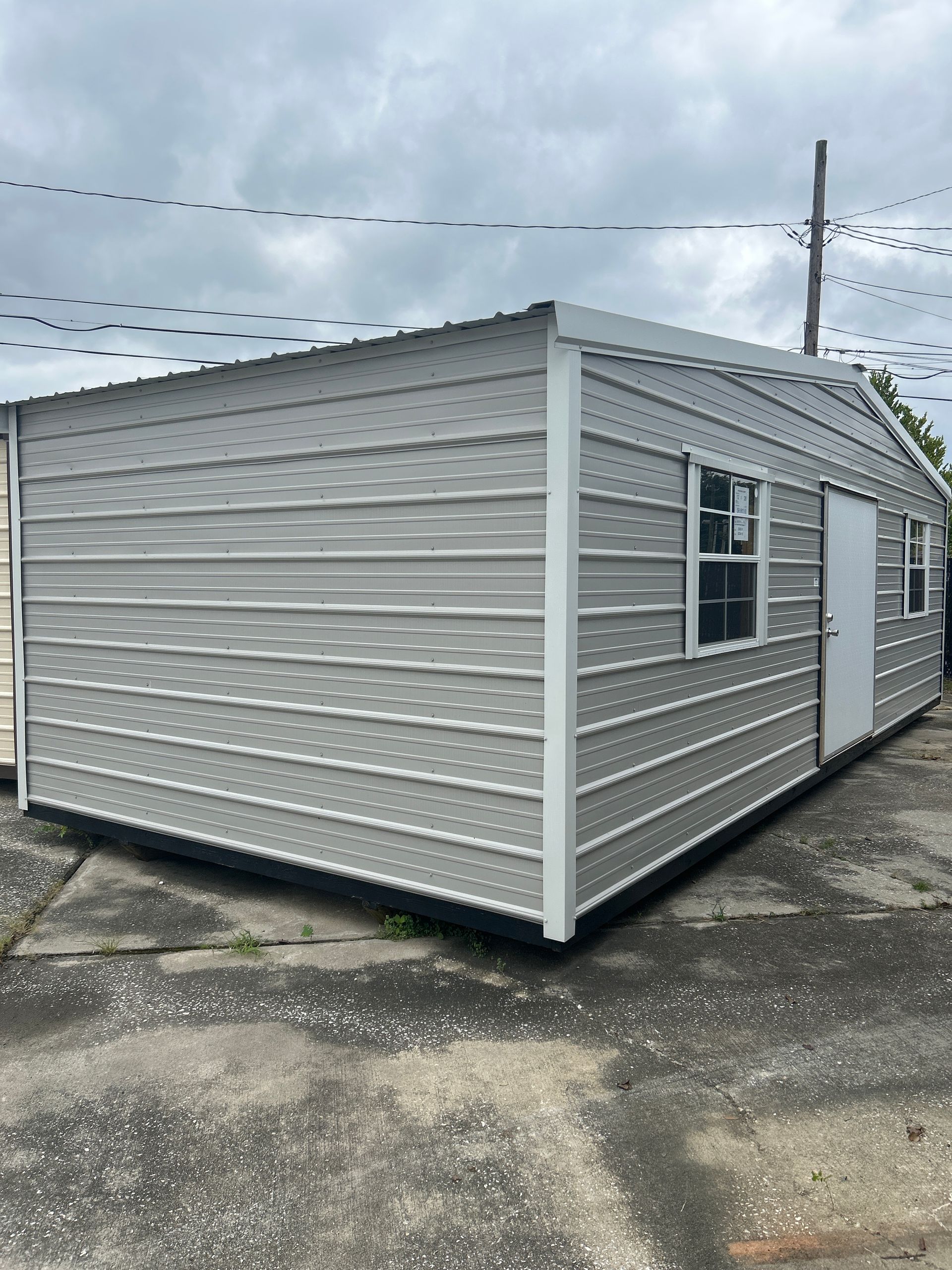 Gray metal storage shed with a white door and two windows, under an overcast sky.