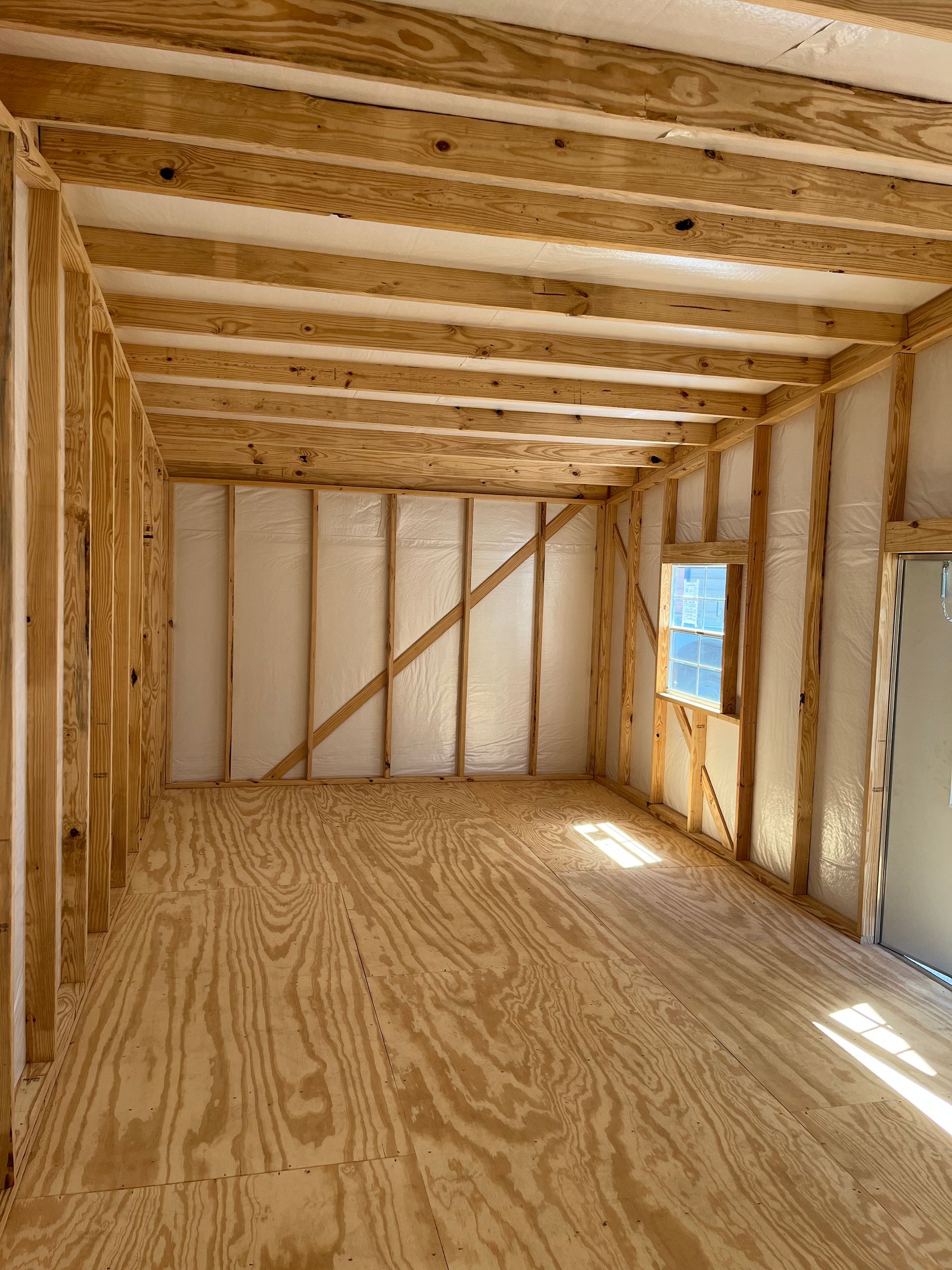 Interior of an unfinished wooden room with exposed framing, plywood floor, and a small window.