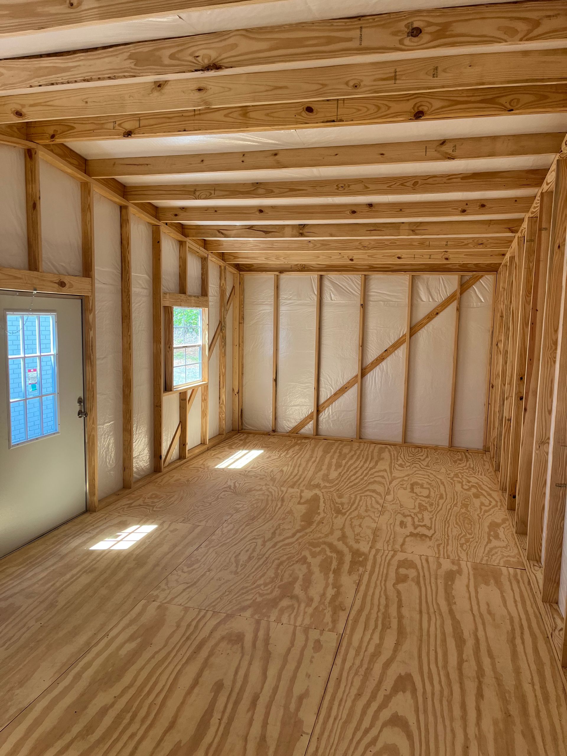 Empty unfinished room with plywood floor, exposed wood framing, and two small windows letting in light