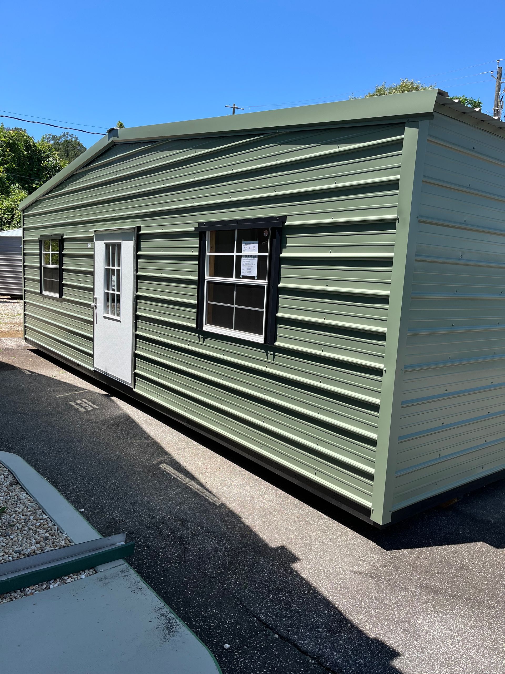 Green corrugated metal shed with white door and black-framed windows on a paved lot under blue sky