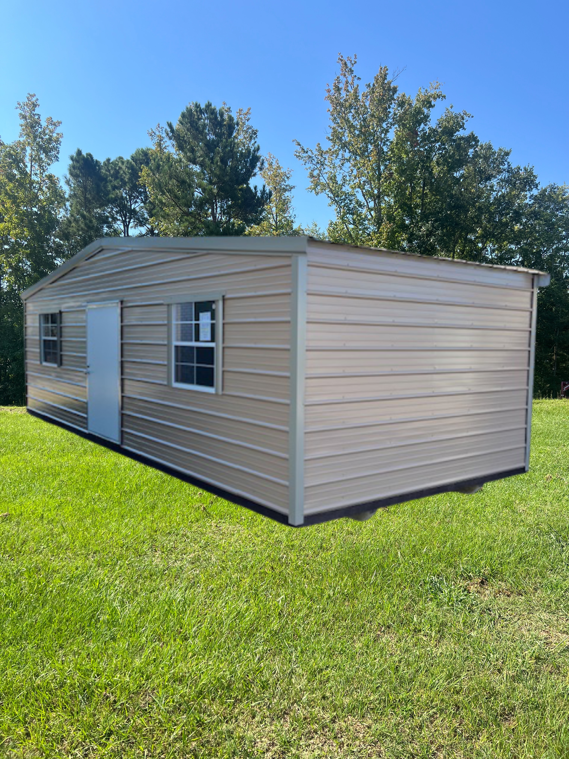 Tan metal shed with door, two windows, set on green grass with trees and blue sky.
