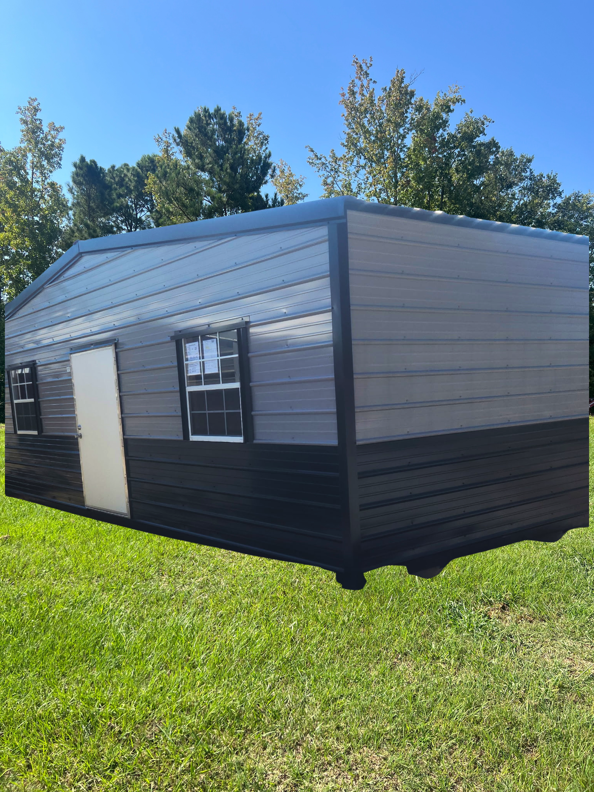 A grey and black rectangular storage shed with a white door and two windows sitting on a grassy lawn under a blue sky.