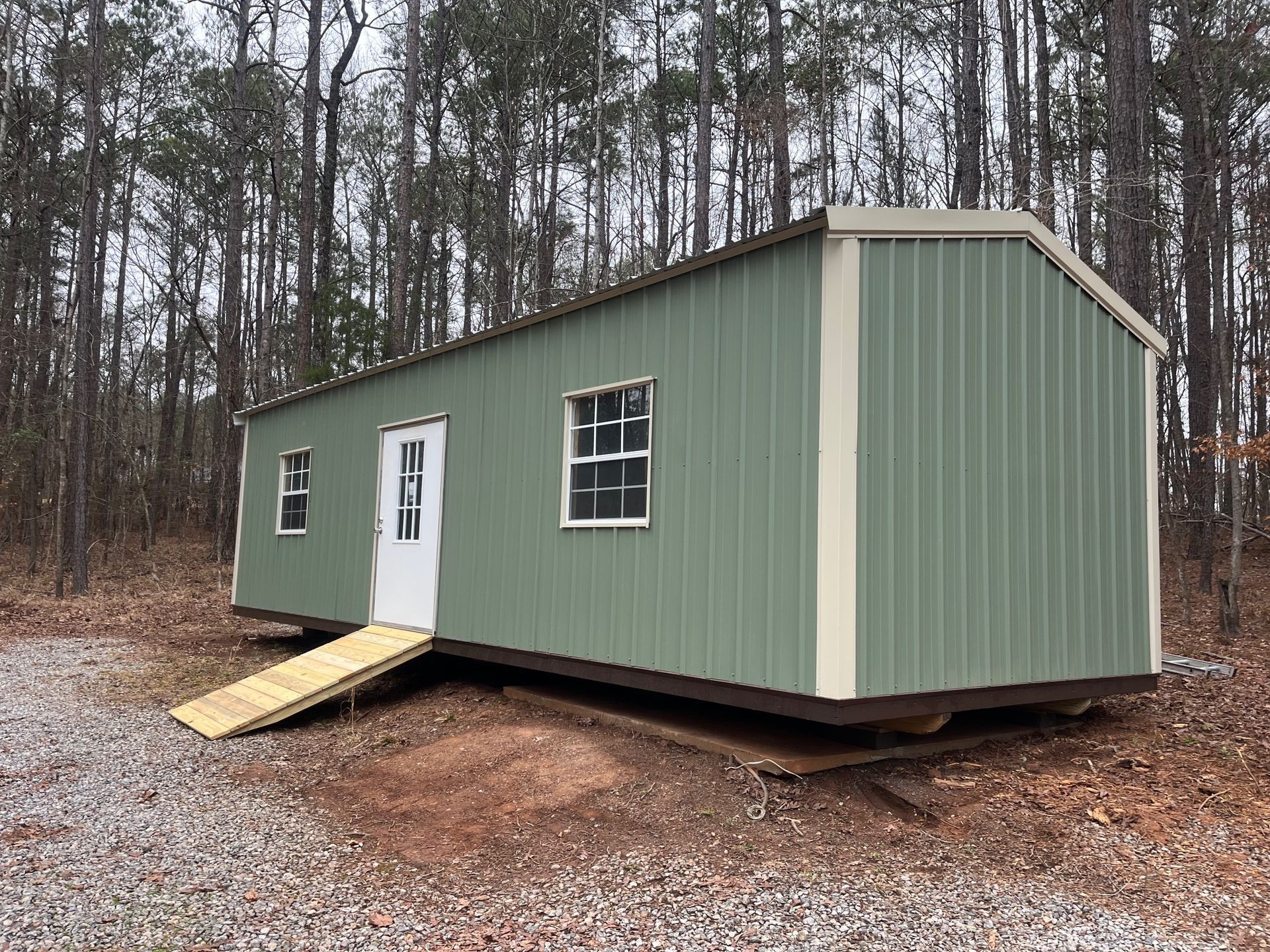 Green metal building with white door, windows, and ramp in a wooded area.