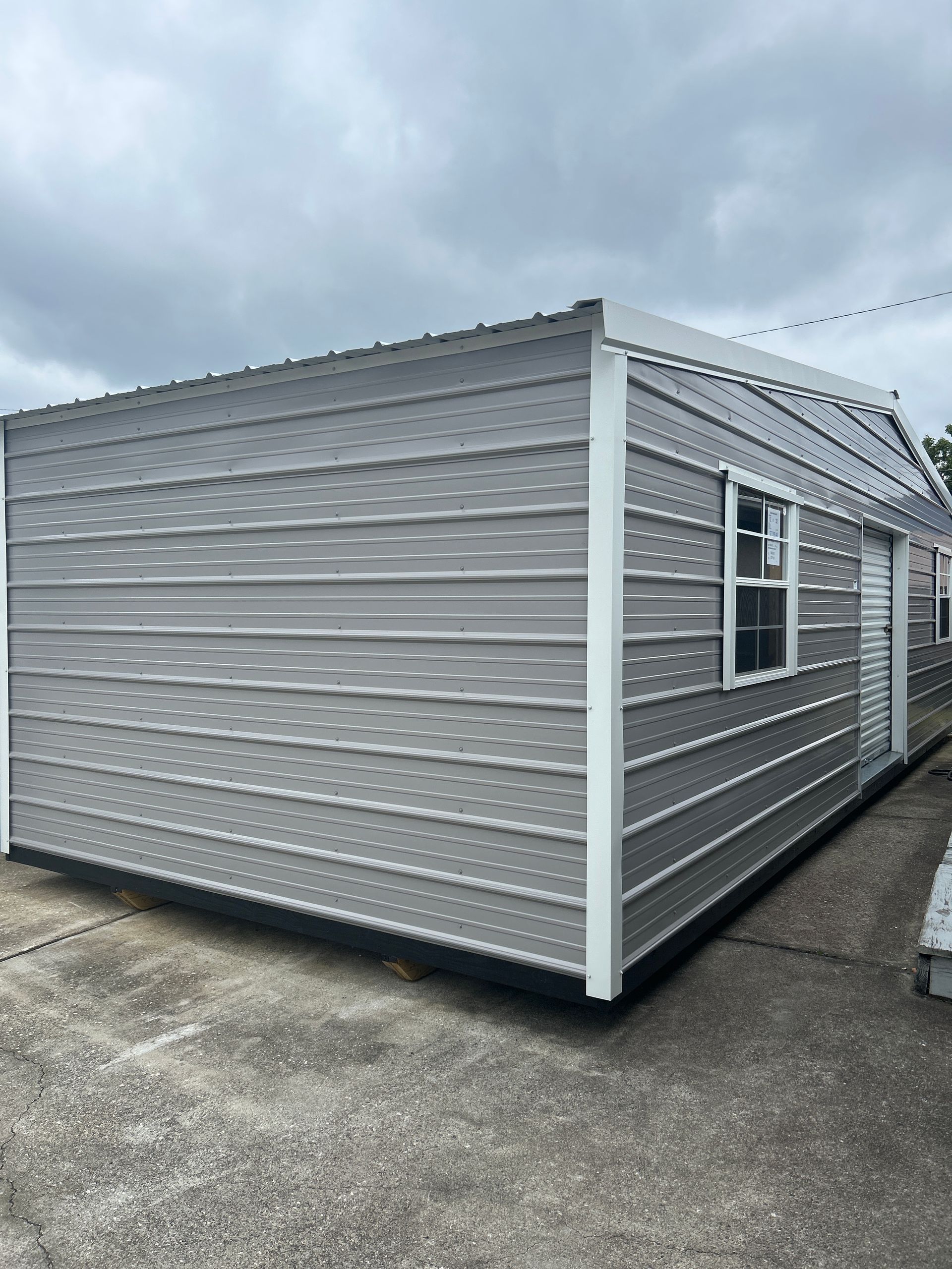 Gray metal building with white trim, windows, and door against a cloudy sky.