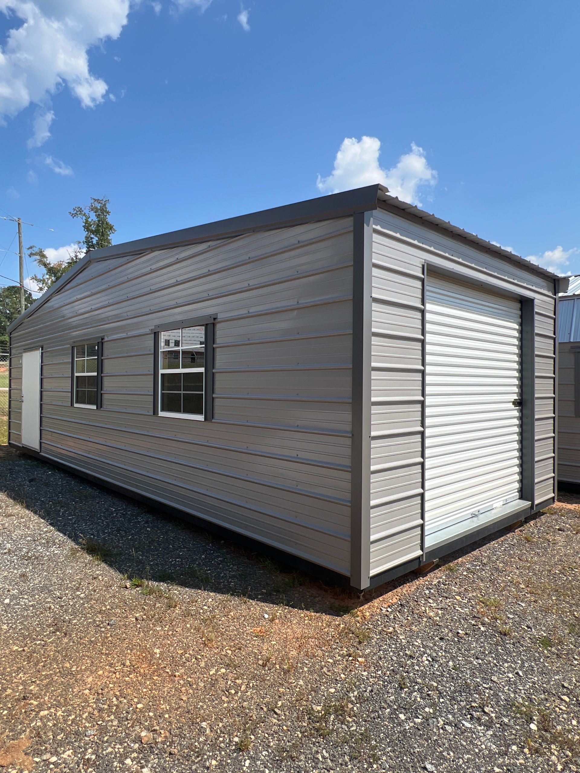 Gray metal shed with a garage door, two windows, and a side door on a gravel lot under a blue sky.