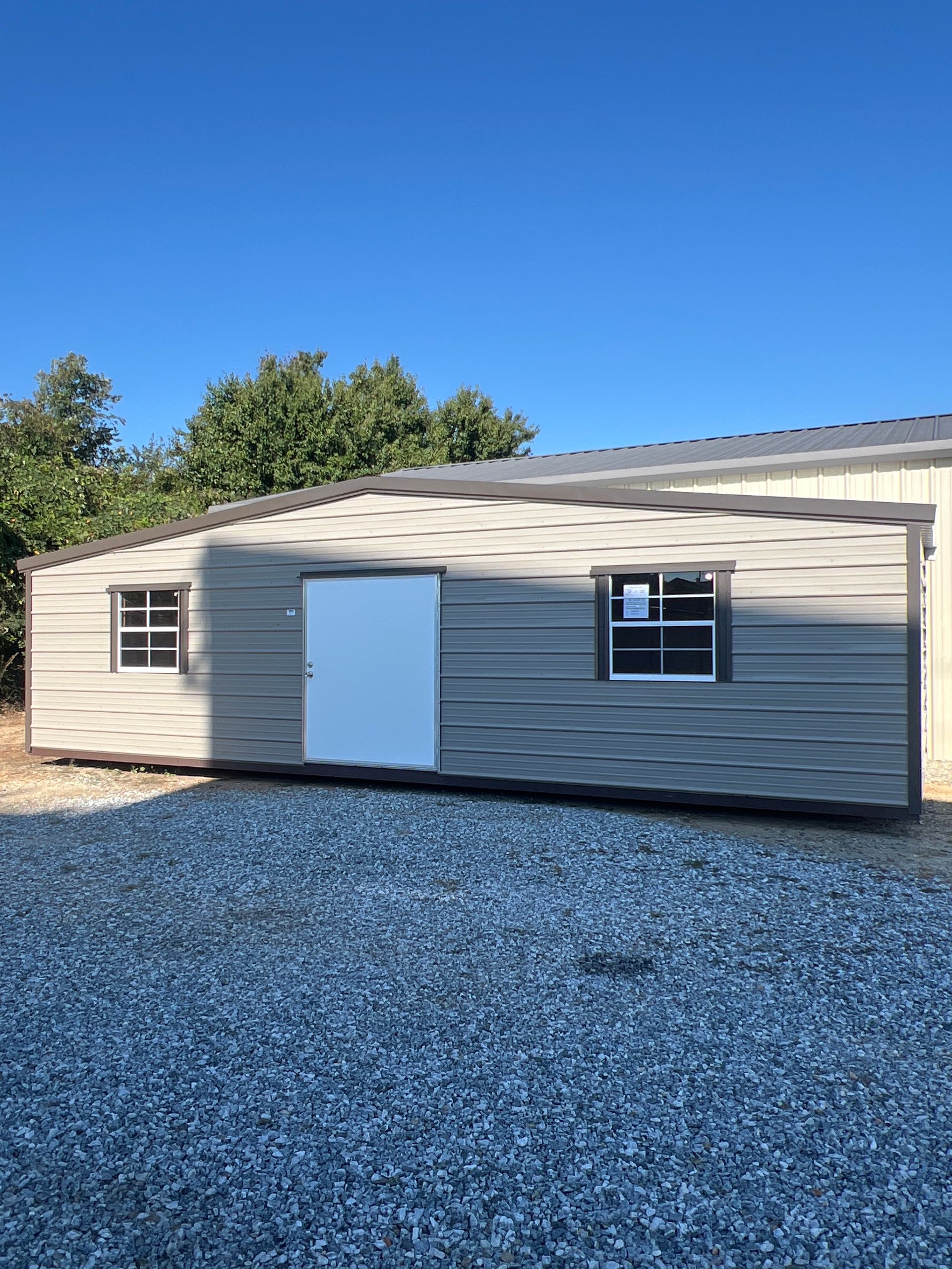 Gray shed with white door and two windows on a gravel surface under a blue sky.