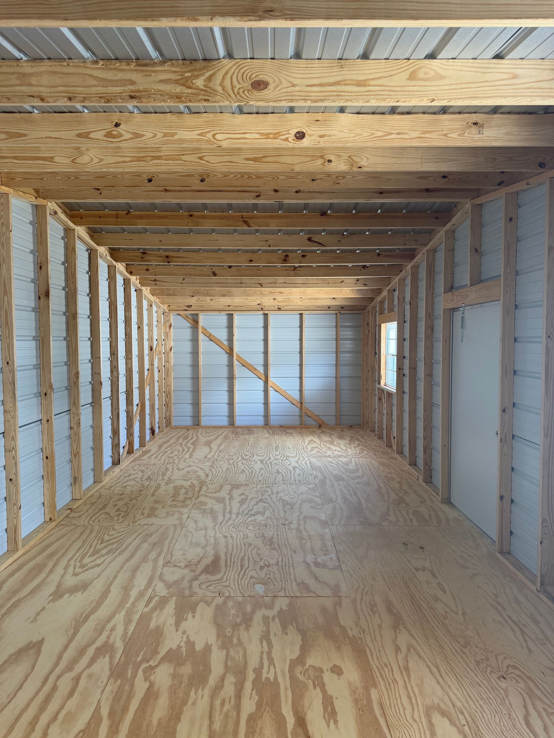 Empty unfinished room with exposed wood framing, plywood floor, and white insulated wall panels.