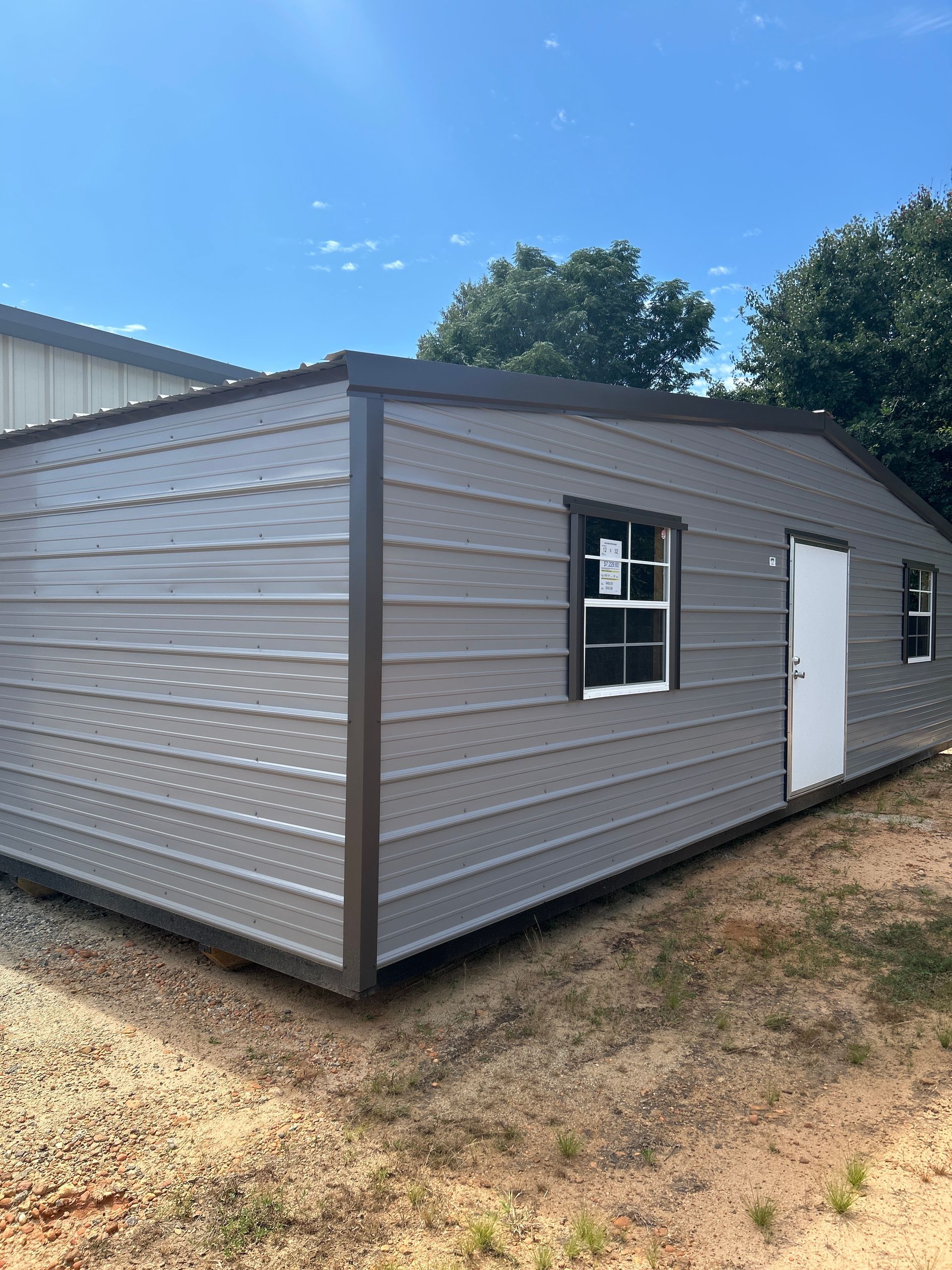 Gray metal shed with brown trim, white door and windows, on a sunny day.
