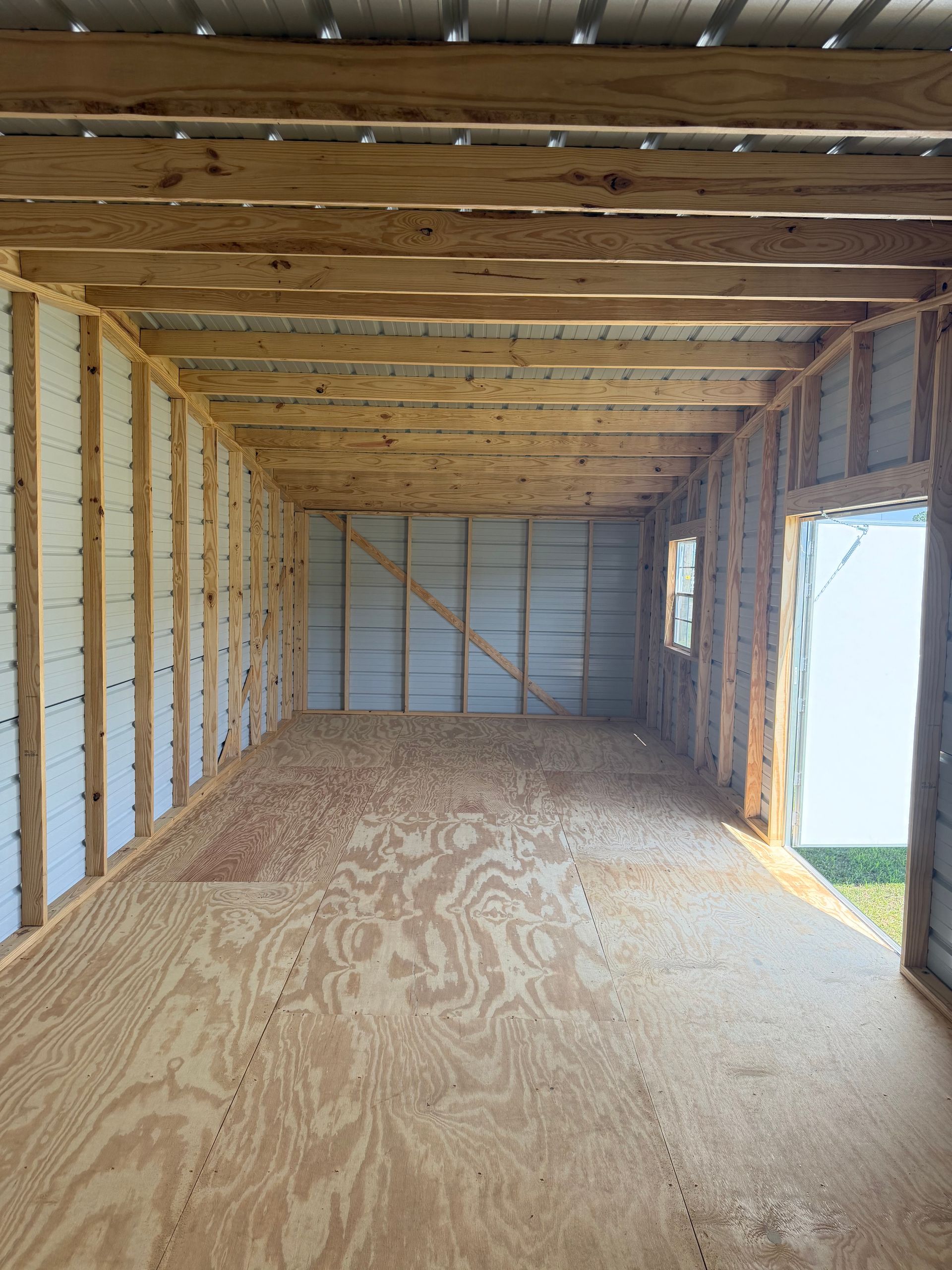 Empty unfinished wooden room with plywood floor, exposed studs, and an open doorway to the outside.