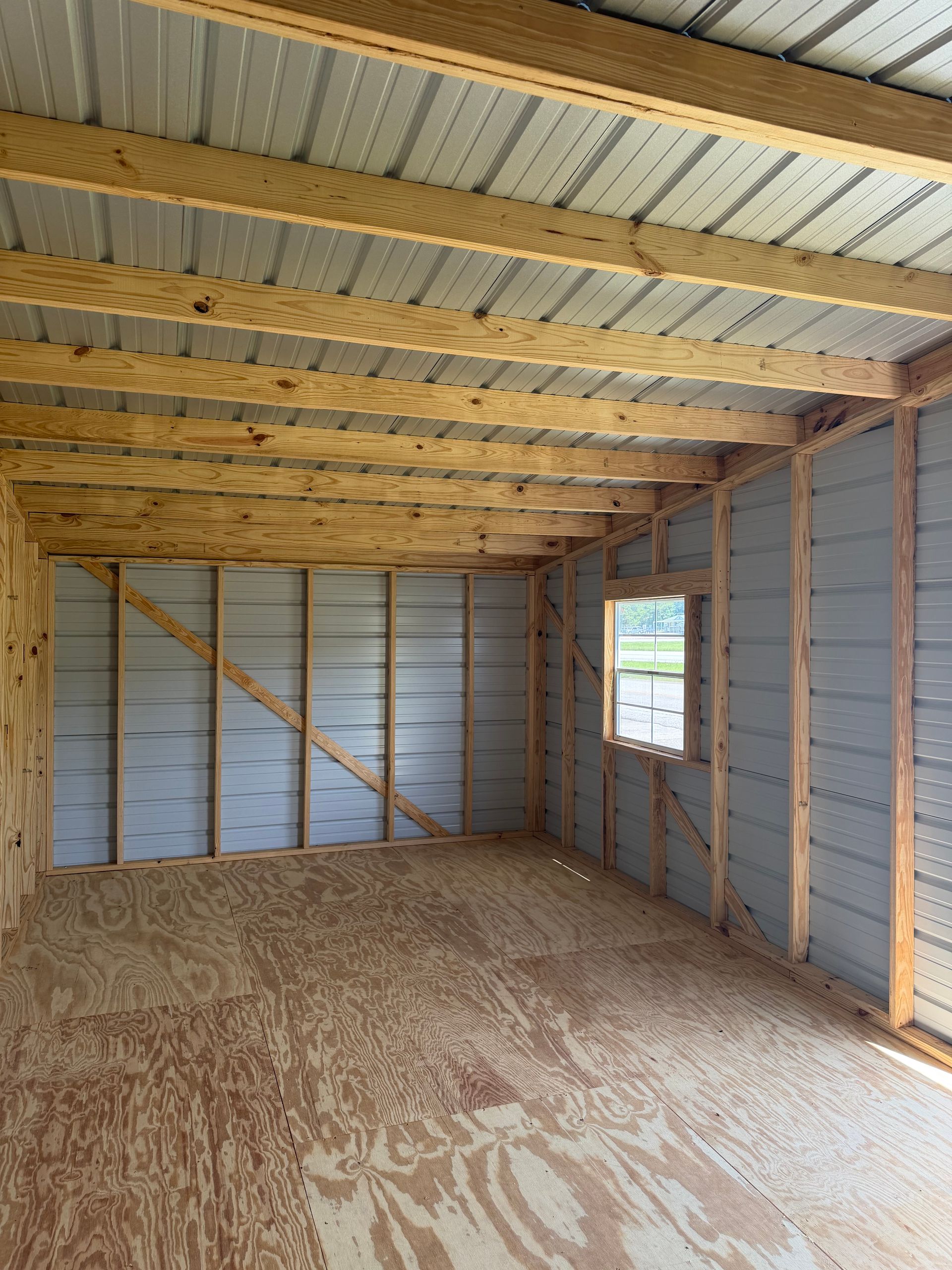 Empty unfinished room with exposed wooden framing, plywood floor, metal walls, and a small window.