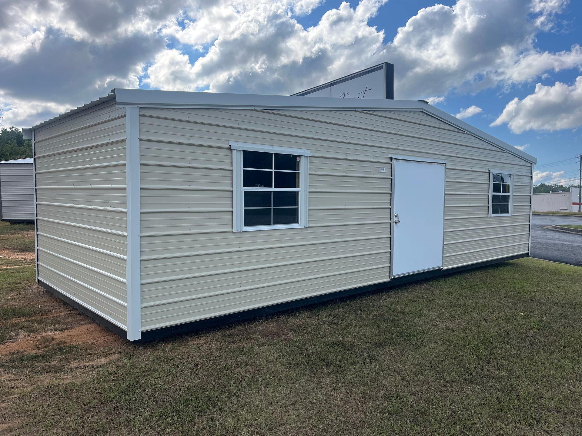 Beige metal building with a white door and two windows on a grassy lot under a cloudy sky.
