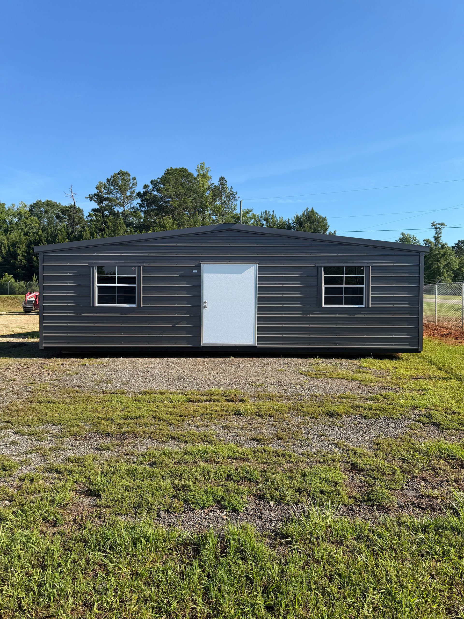 Small dark gray mobile home with white door and windows in a grassy field under a clear blue sky