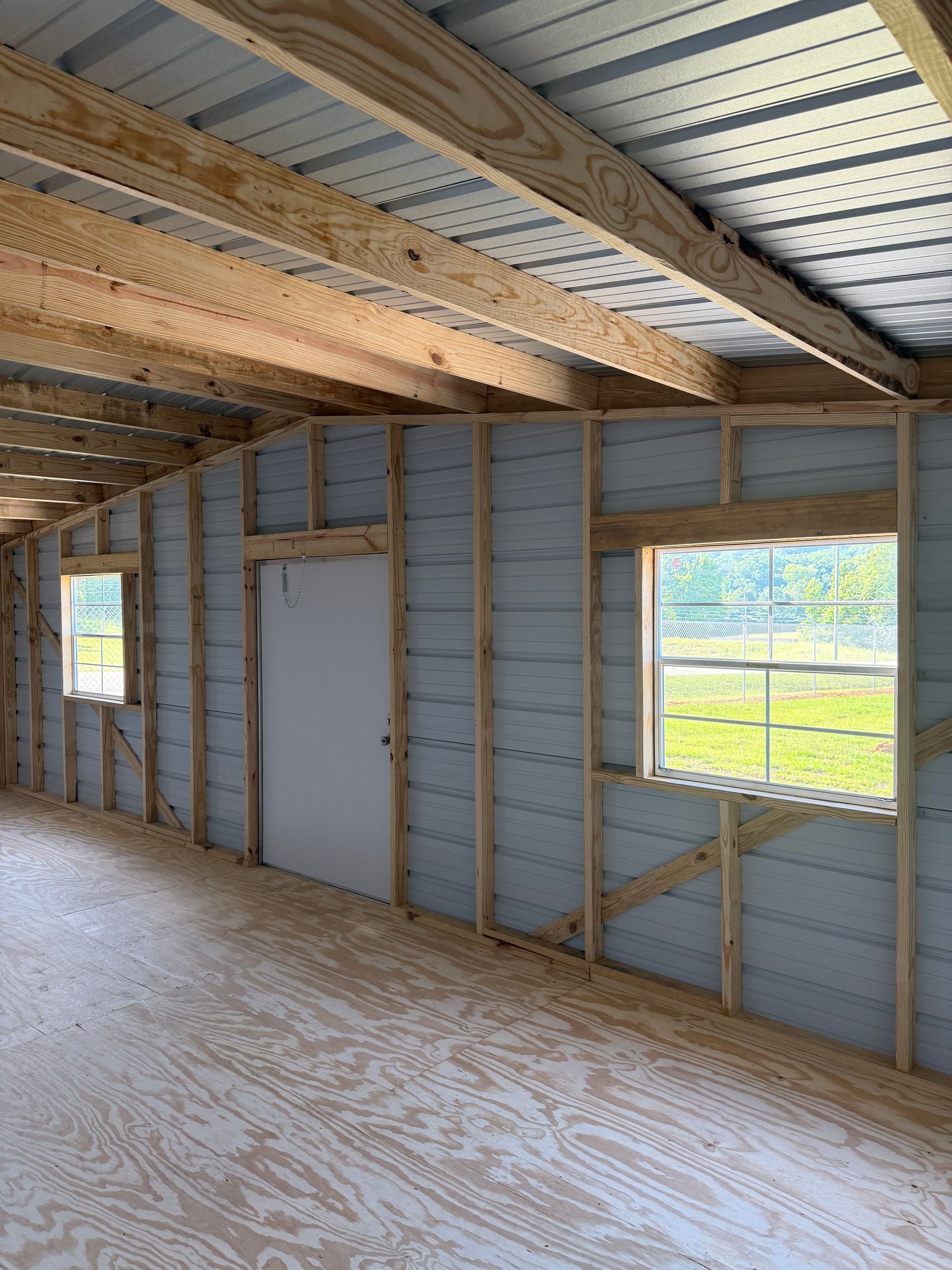 Interior of an unfinished room with exposed framing, insulation, a plywood floor, and two windows.
