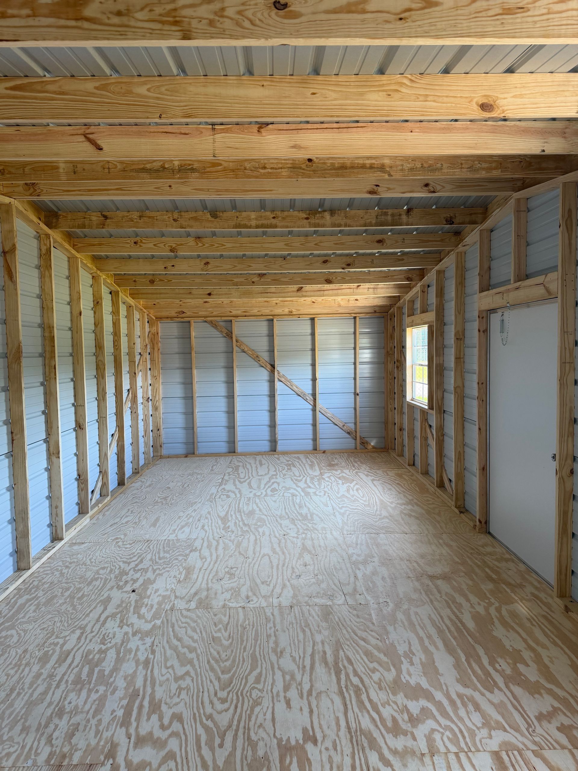 Empty unfinished room with plywood floor, exposed wall studs, and ceiling joists, with a small window and door.