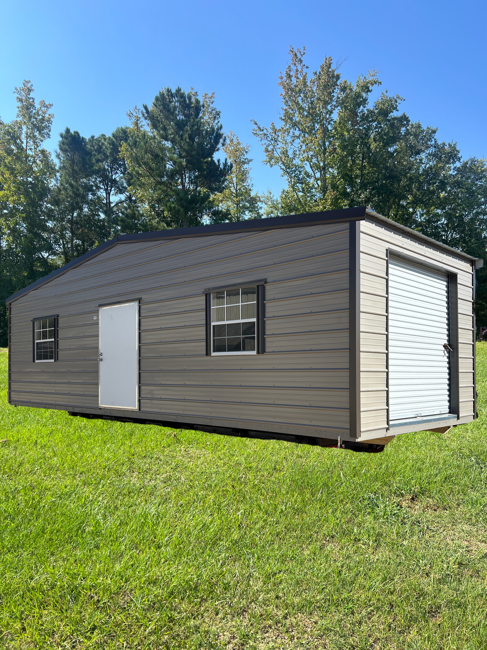 Gray and brown metal shed with garage door, windows, and a door on a grassy lawn.