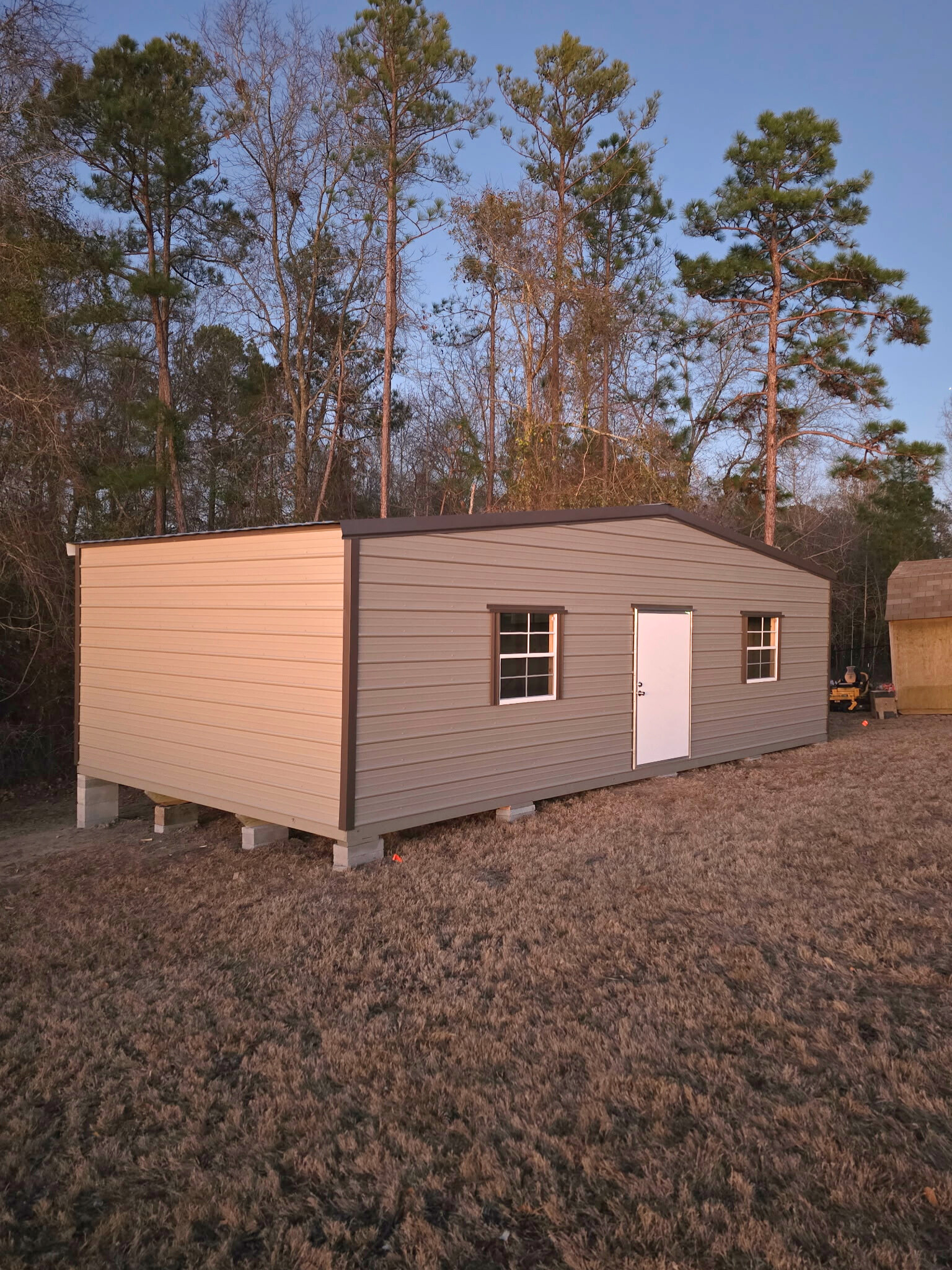A small, gray and tan shed with a white door and windows sits in a grassy area with trees behind it.
