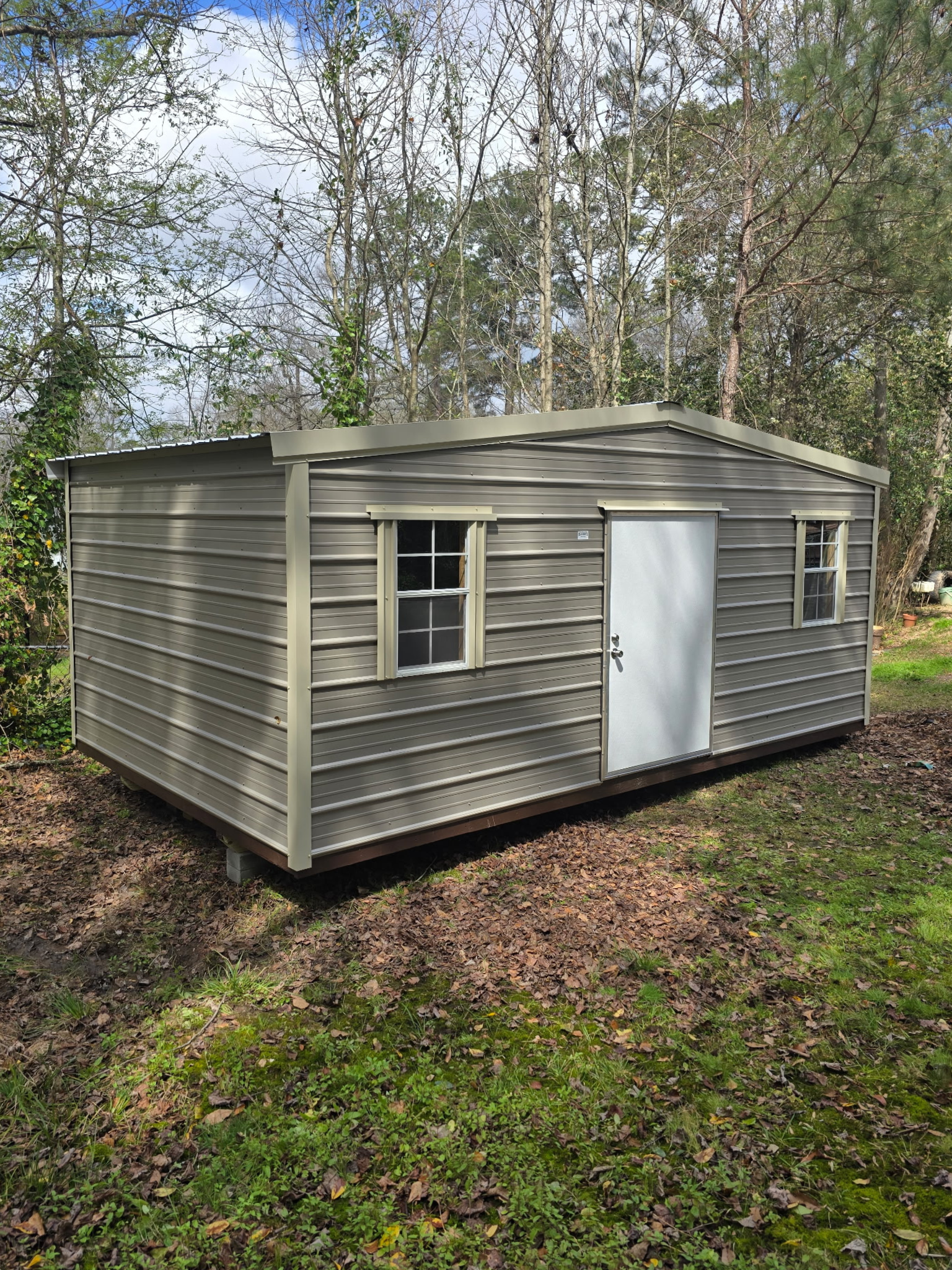 Tan shed with windows and a door in a grassy area with trees.