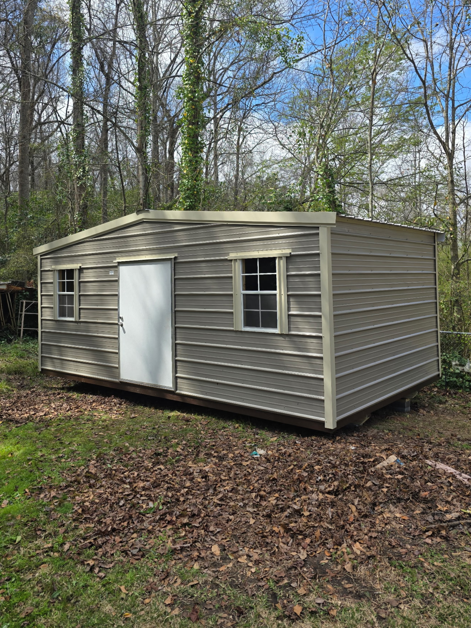 Tan metal shed with white door and windows, surrounded by trees and leaves.