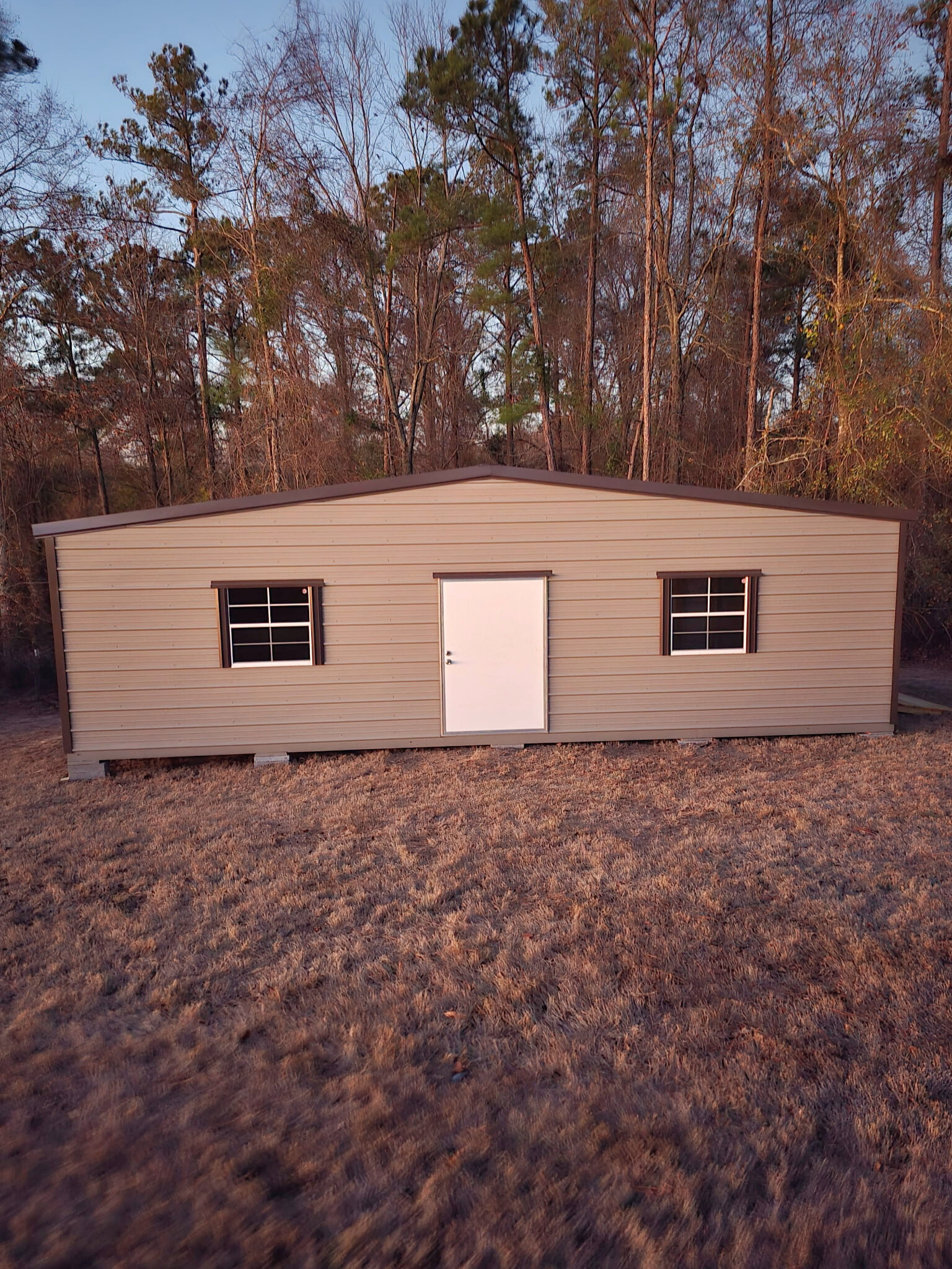 Tan shed with a white door and two windows sits in a grassy area with trees in the background.