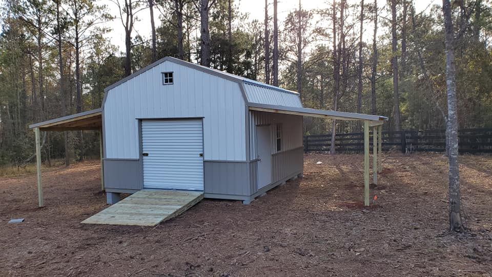 Barn-style shed with ramp and two canopies, white and gray siding, in a wooded outdoor setting.