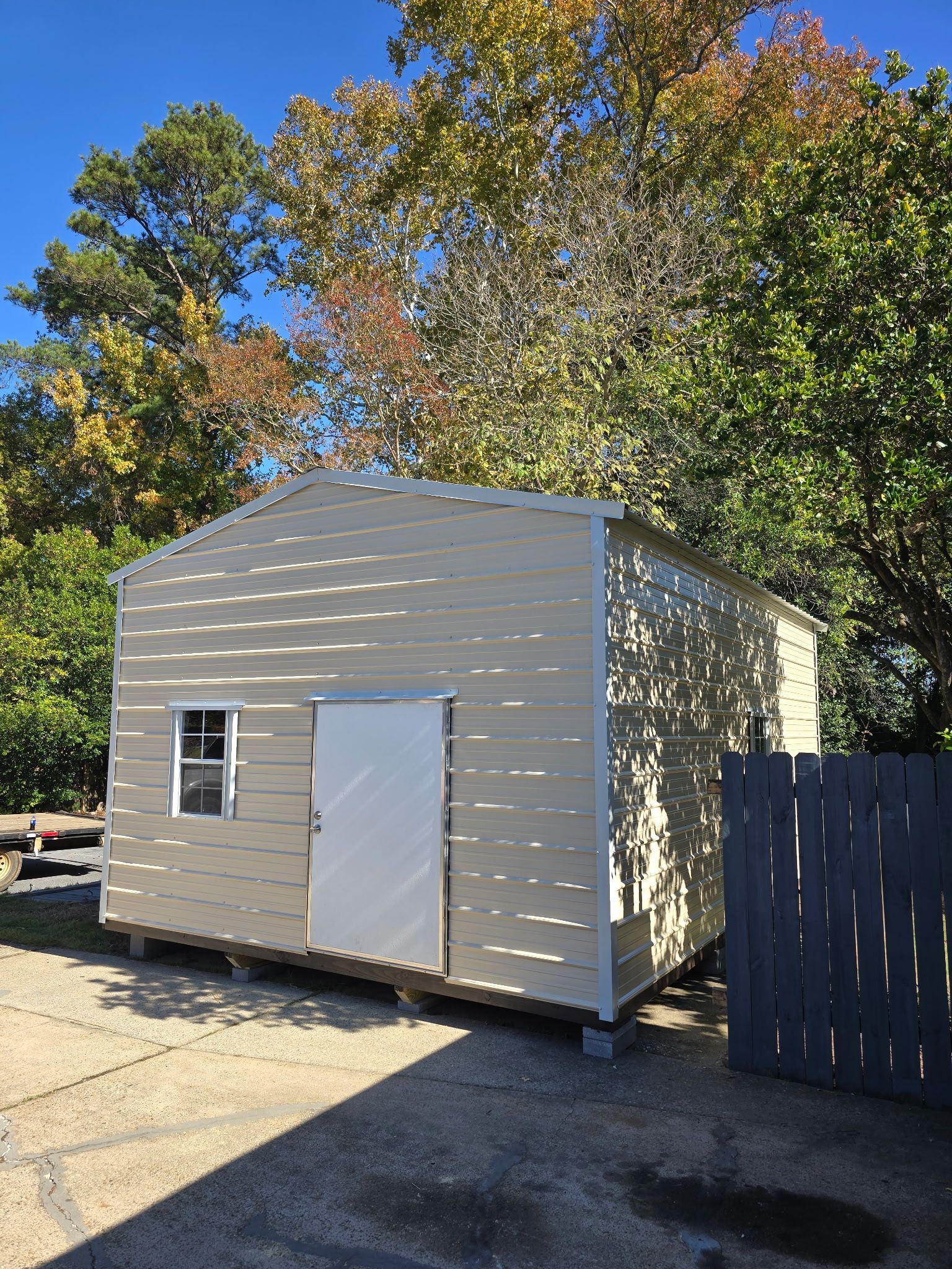 Beige metal shed with a white door and small window, set outside with fall foliage in background.