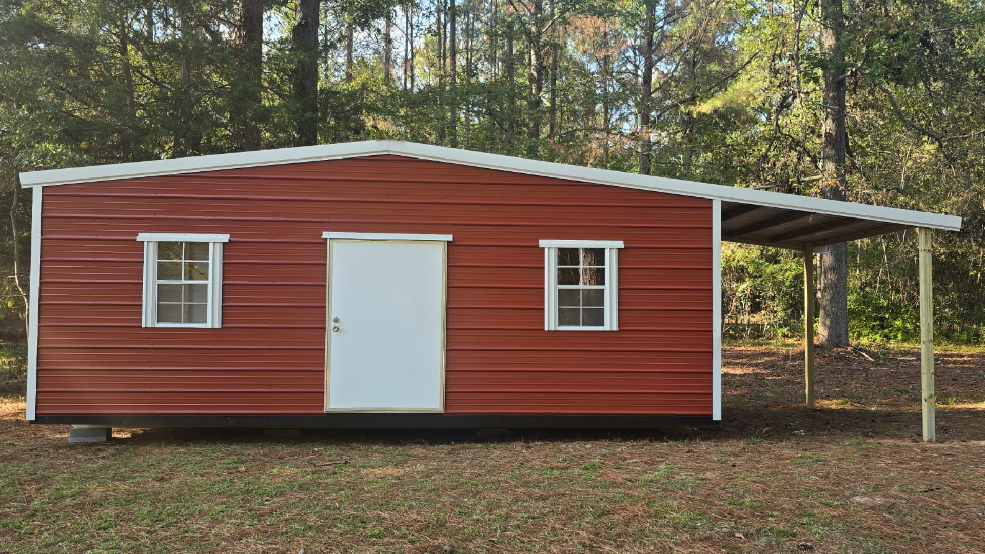 Red metal building with white trim, windows, and door. Carport attached on the right.