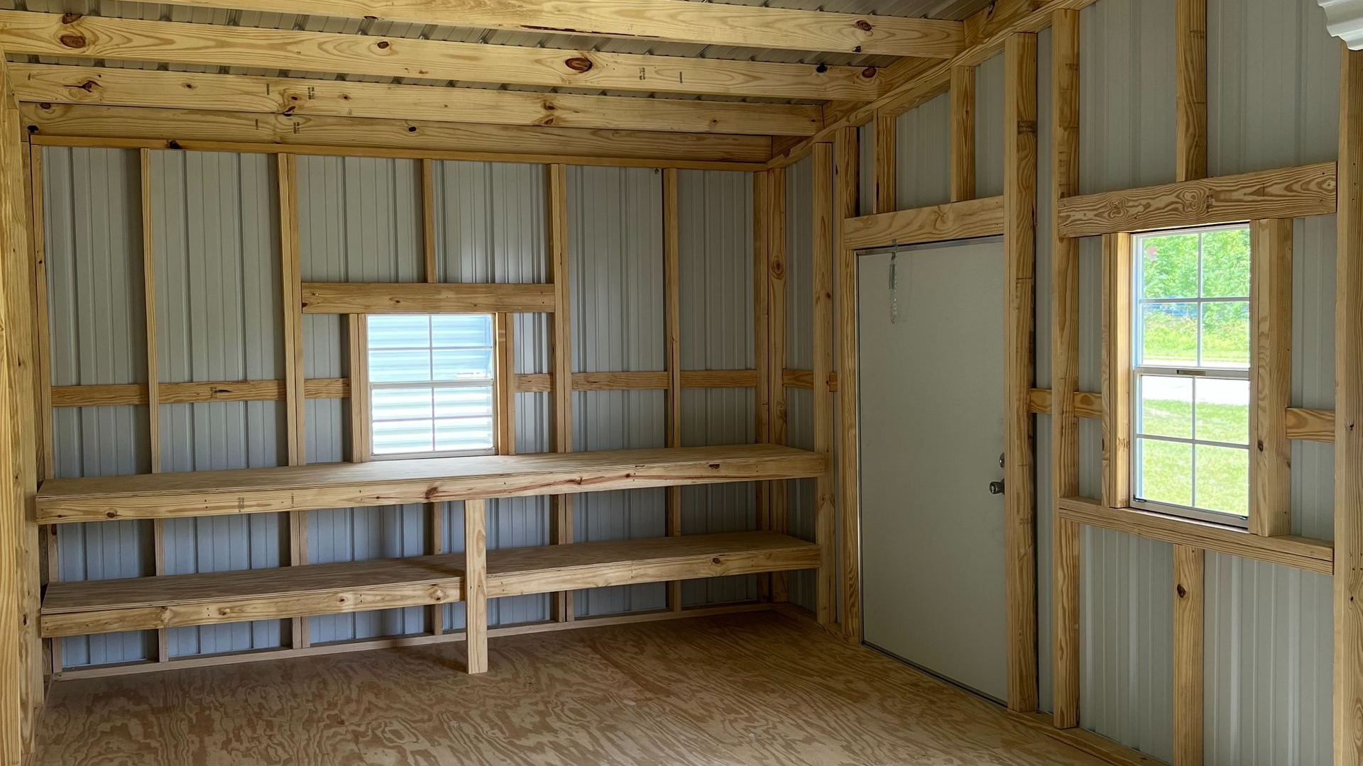 Interior of a shed with wood framing, metal siding, two windows, door, and built-in shelves.