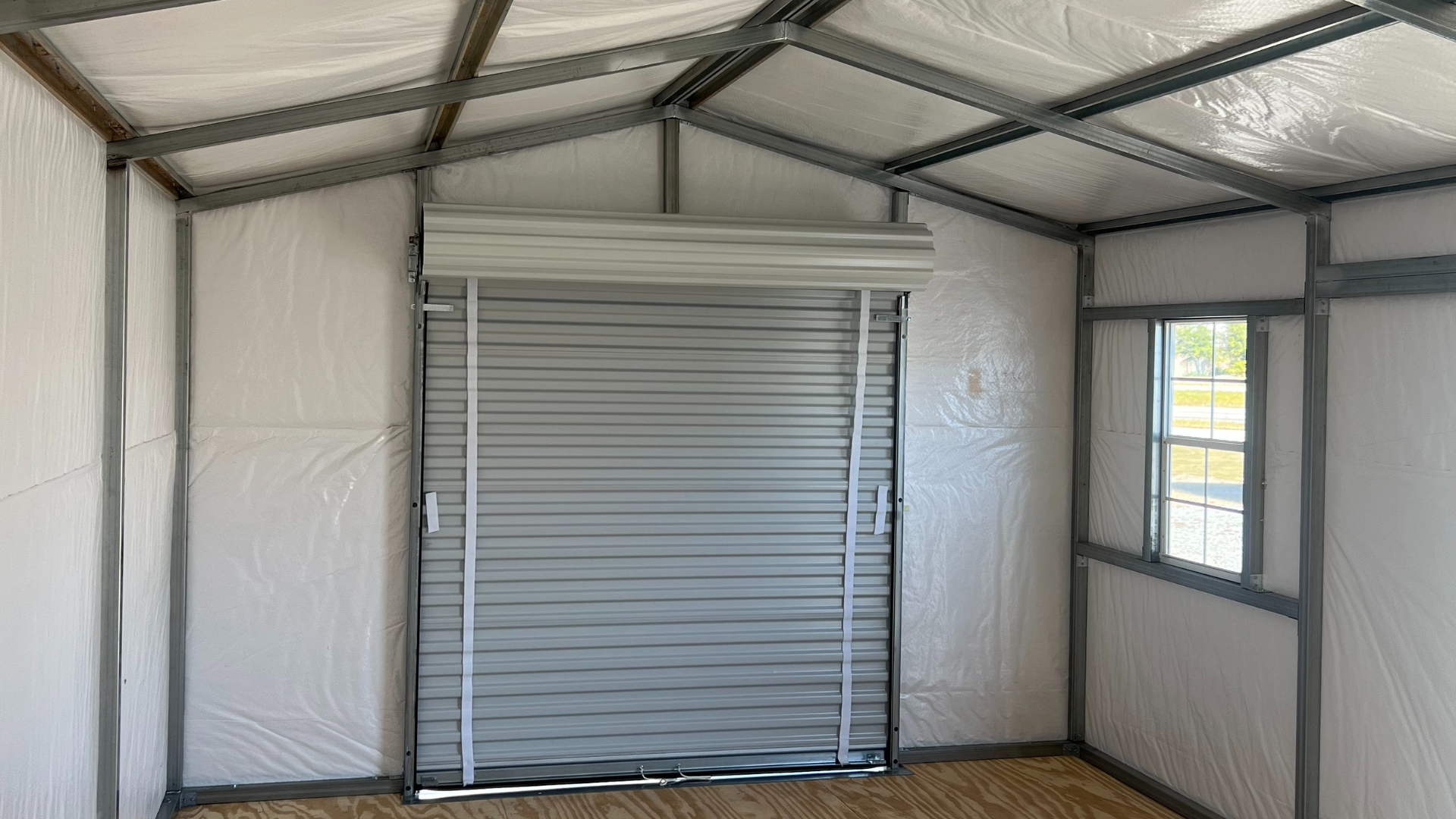 Interior of a metal shed with a closed gray roll-up door, window, and white interior walls.